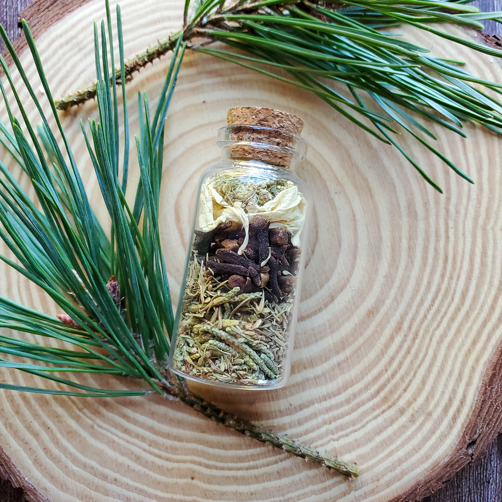 Small glass bottle with dried herbs on a wooden surface with pine branches. Abundance Spell Bottle, Intention Setting Herbs for Prosperity and Sucess