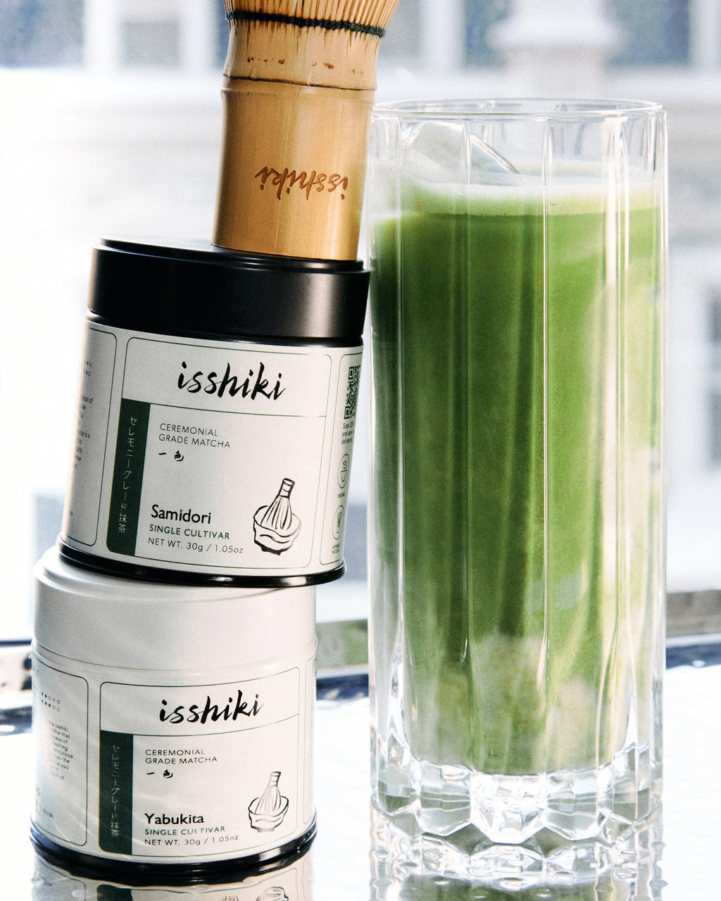Container of 'isshiki' matcha with a glass of green tea and a bamboo whisk on a blurred background