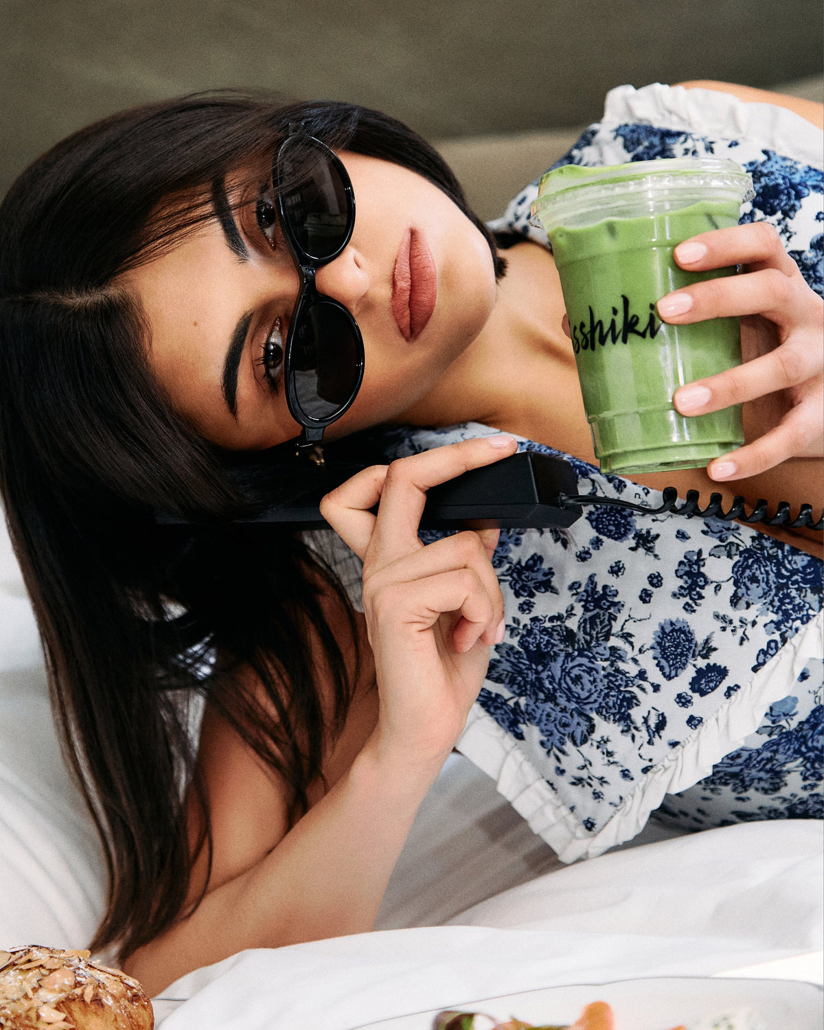 Woman lying down with a green smoothie and phone, wearing sunglasses.
