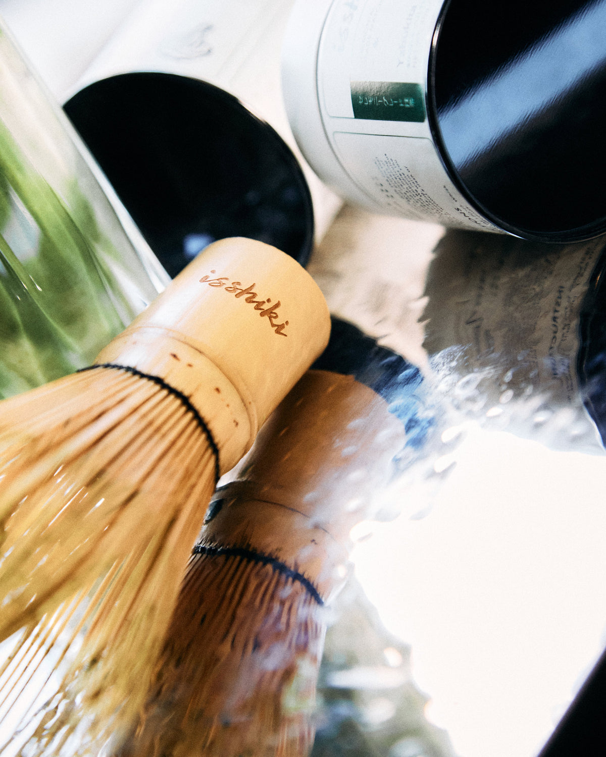 Matcha whisk and container on a reflective surface with greenery in the background