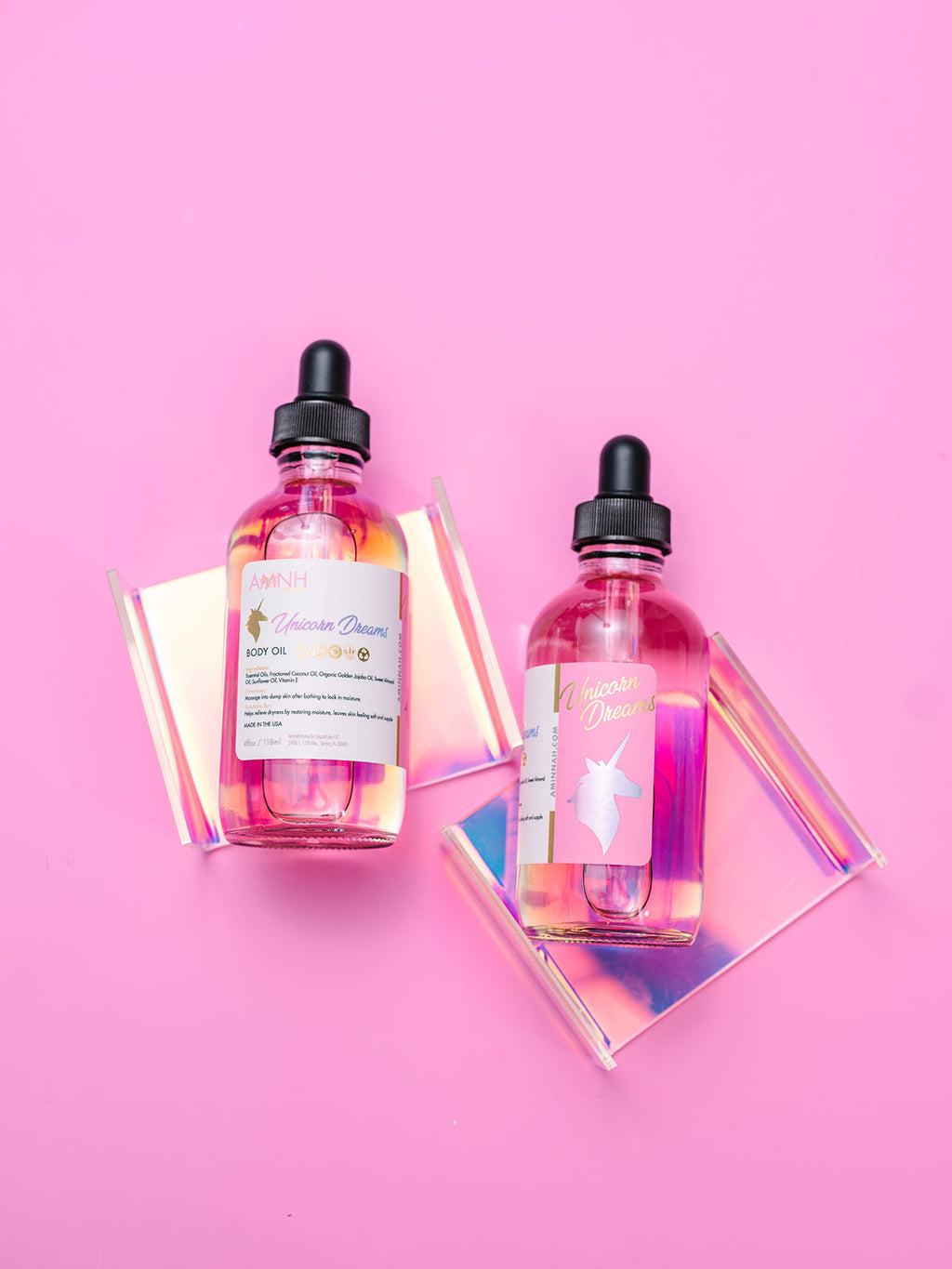 AMINNAH Unicorn Dreams Body Oil displayed in two glass dropper bottles on a soft pink background with iridescent acrylic accents. This clean beauty, cruelty-free, vegan body oil is formulated with naturally derived jojoba oil, coconut oil, rosehip oil, hazelnut oil, avocado oil, sunflower oil, and vitamin E to deeply hydrate, nourish, and soften skin while leaving a silky, radiant glow. Infused with a sweet cotton candy-inspired fragrance, this female-owned brand is available at Tree of Life Grocers.