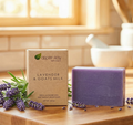 A bar of lavender and goats milk soap stands next to its kraft paper packaging box on a wooden table, styled with fresh lavender sprigs and rosemary in a warm, sunlit kitchen setting.