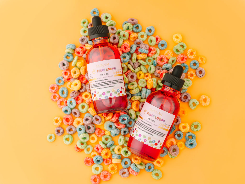 Frut Loops Body Oil displayed with colorful fruity cereal on a yellow background by a female-owned brand. Clean beauty, cruelty-free, vegan body oil made with natural ingredients including niacinamide, licorice extract, and vitamin C to nourish, hydrate, brighten, and leave skin glowing.