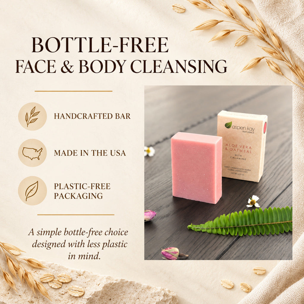 A handcrafted pink face and body cleansing bar shown next to its beige paper packaging on a dark wood surface, surrounded by decorative wheat stalks, a green fern, and small flowers. Text on the left highlights key selling points: handcrafted, made in the USA, and plastic-free packaging.