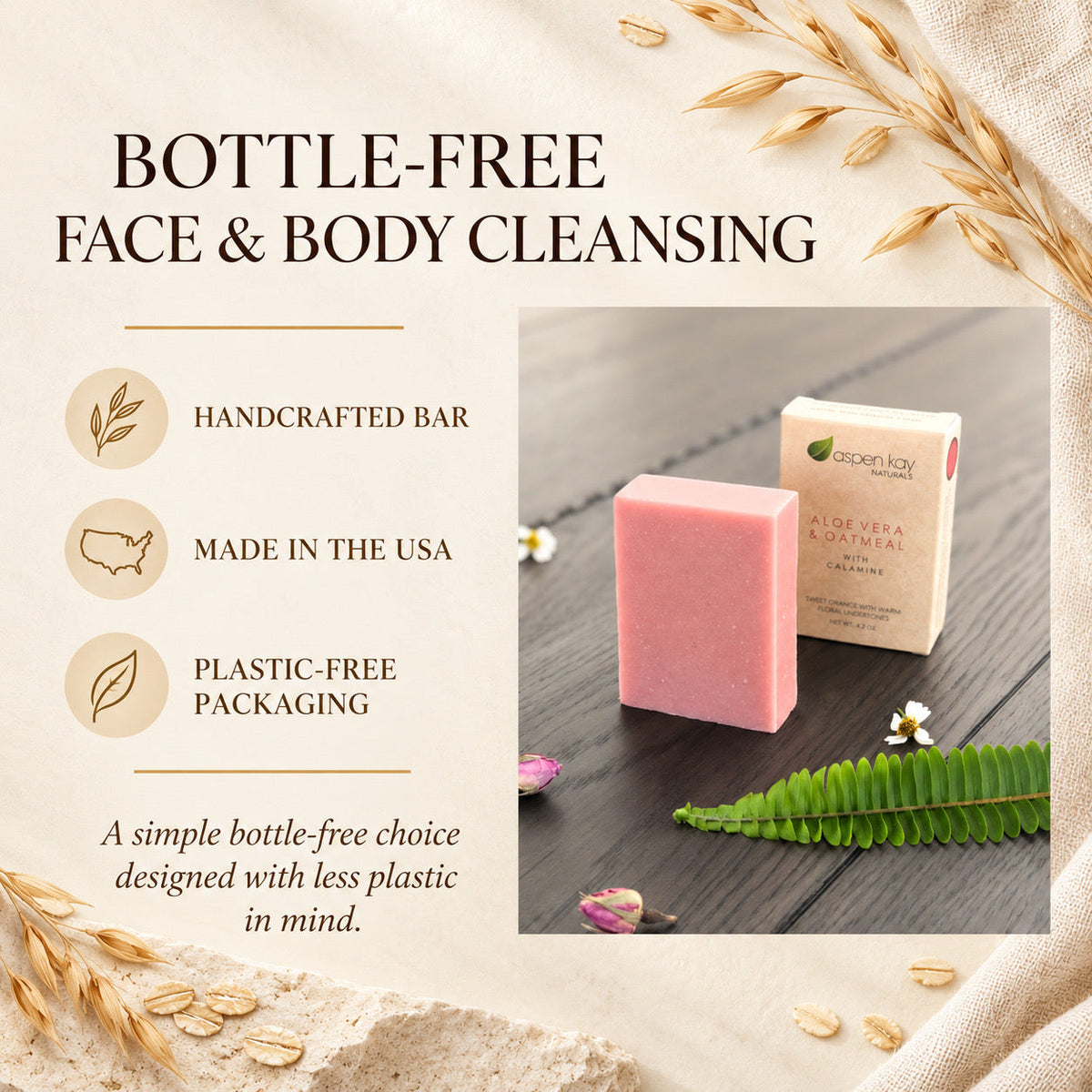 A handcrafted pink face and body cleansing bar shown next to its beige paper packaging on a dark wood surface, surrounded by decorative wheat stalks, a green fern, and small flowers. Text on the left highlights key selling points: handcrafted, made in the USA, and plastic-free packaging.