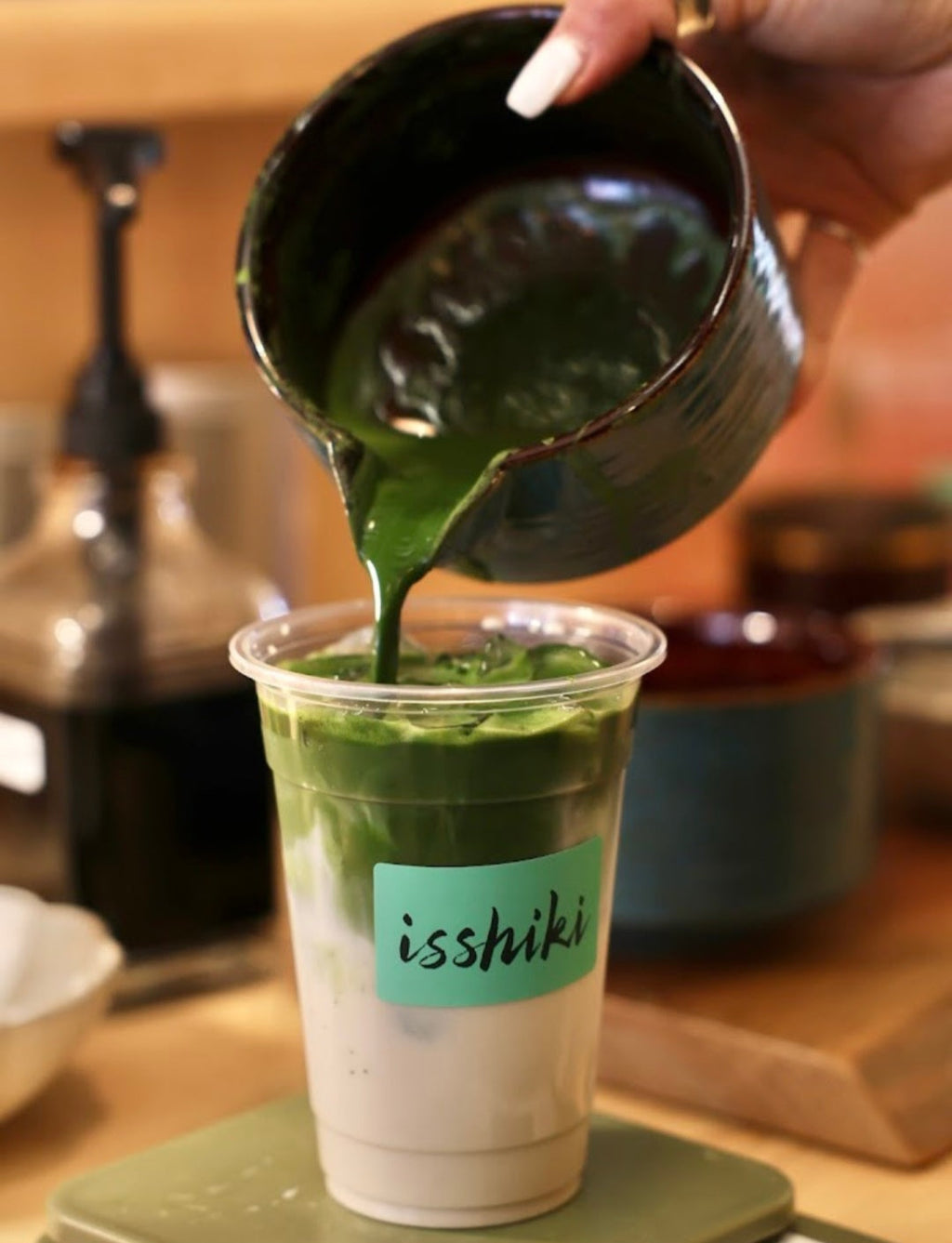 Green liquid being poured from a black cup into a clear cup with 'isshikii' branding on a wooden surface.