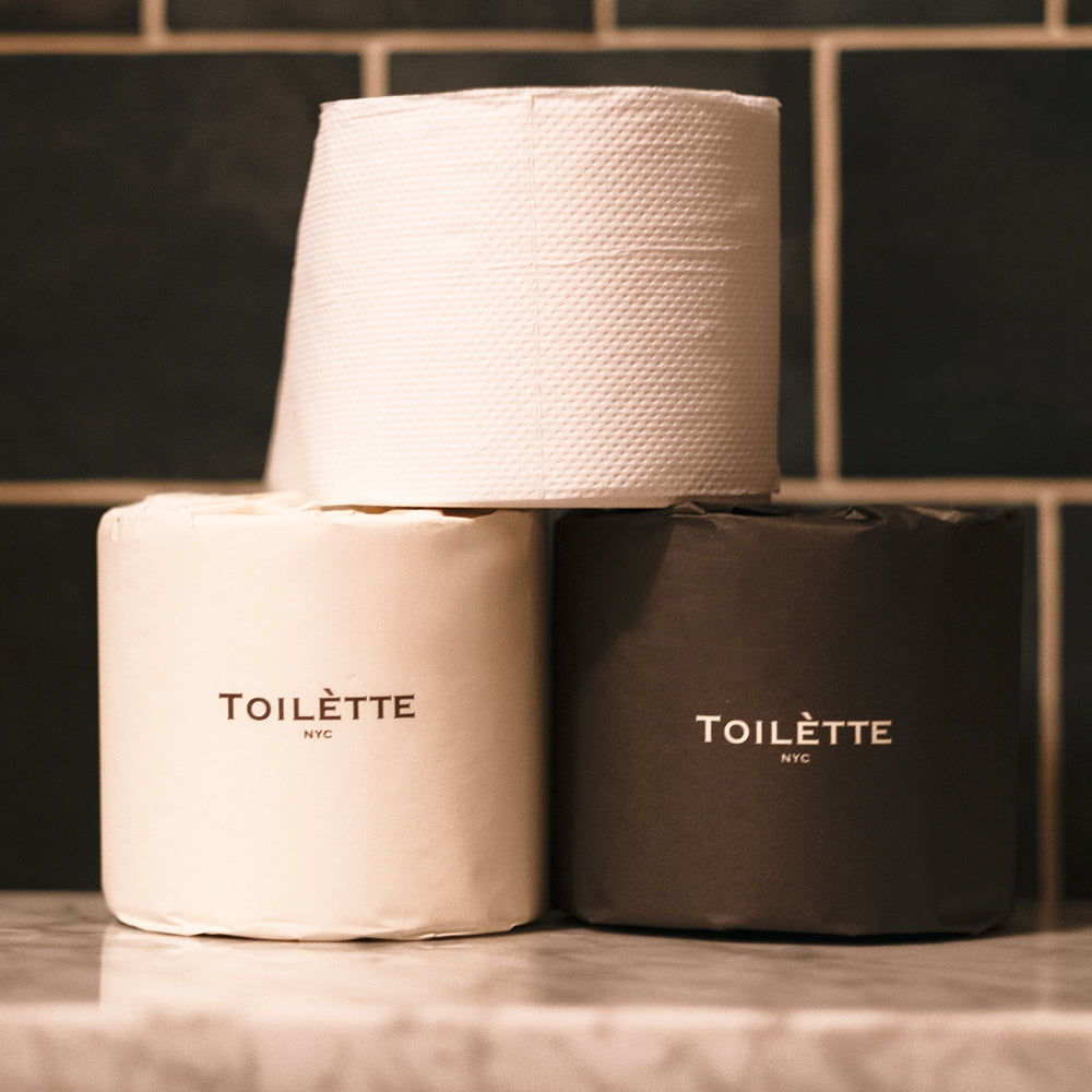 Three rolls of Toilette toilet paper stacked on a marble surface against a dark tiled wall. One roll is off-white, and two are dark charcoal, each printed with the brand name 'TOILLETTE NYC'. 100% Premium Bamboo Toilètte Rolls: Tree-free, lab-tested and 100% free of PFAS, formaldehyde, and elemental chlorine.