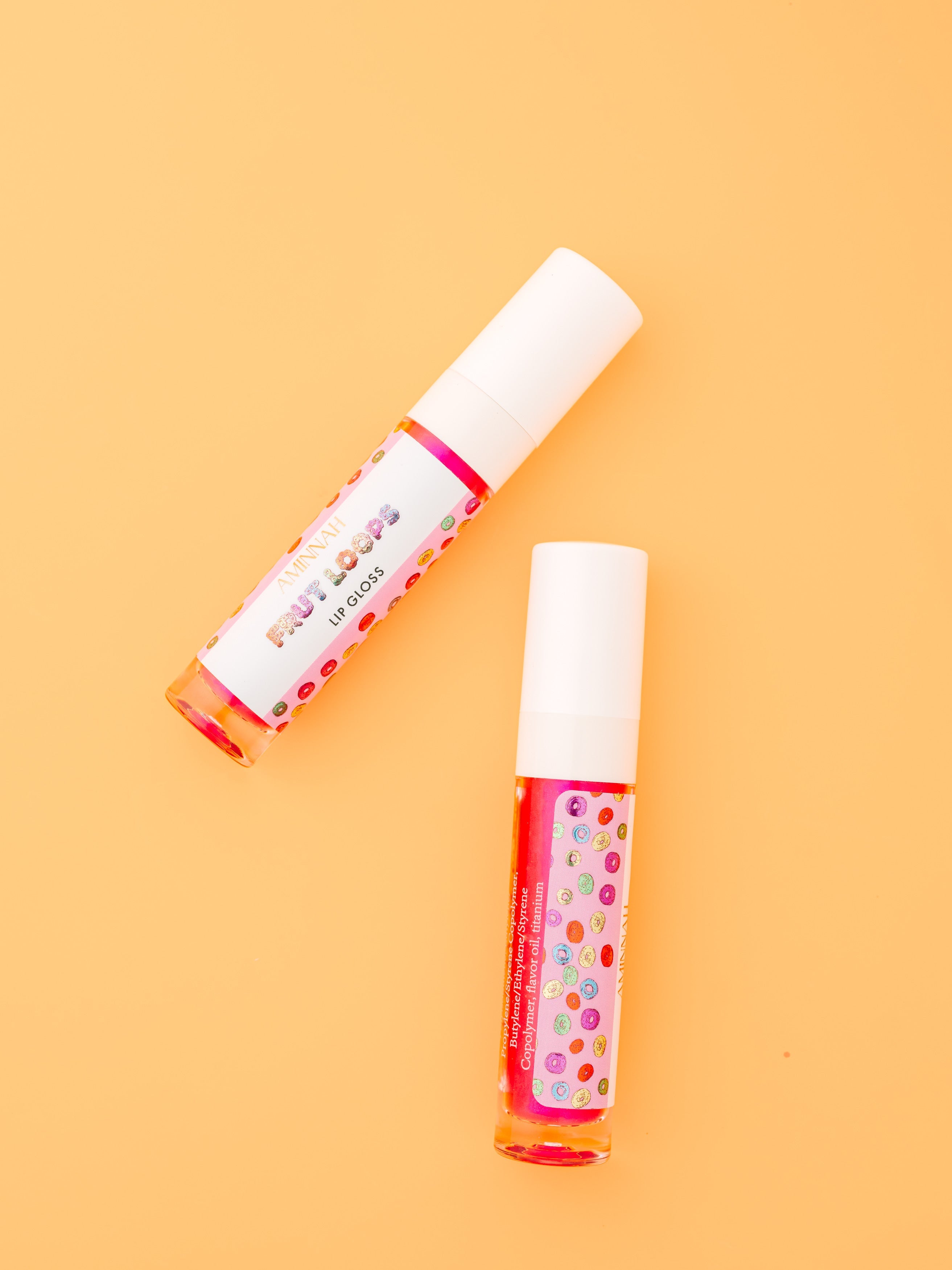 Two tubes of AMINNAH Frut Loops Lip Gloss displayed on a soft peach background, featuring a fruity cereal-inspired design. High-shine moisturizing vegan lip gloss made with coconut oil and naturally derived ingredients. Clean beauty, cruelty-free, female-owned brand, made in the USA, available at Tree of Life Grocers.