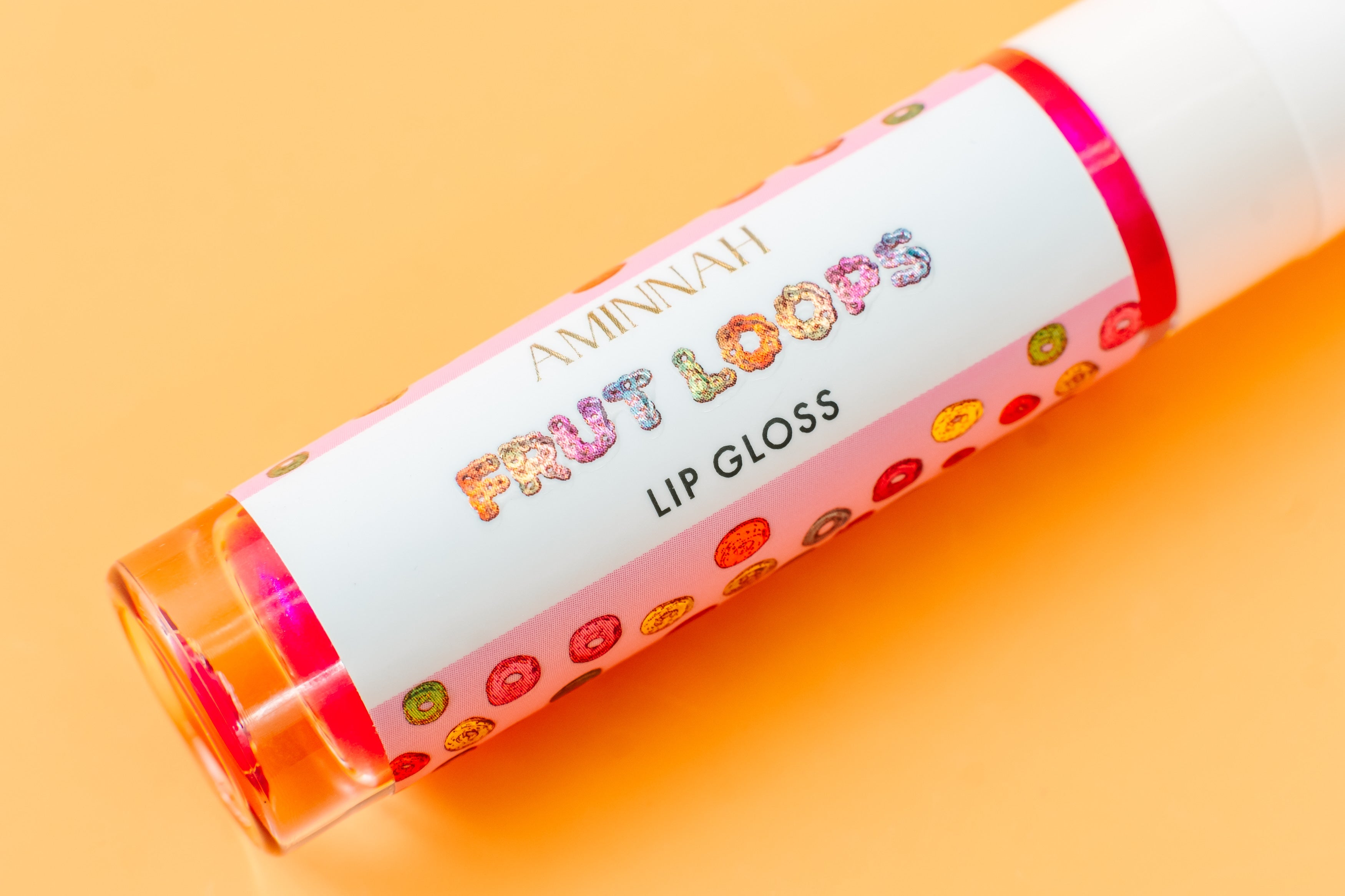 Close-up of AMINNAH Frut Loops Lip Gloss tube featuring colorful fruity cereal-inspired packaging on a warm peach background. Moisturizing high-shine vegan lip gloss made with coconut oil and naturally derived ingredients for soft, hydrated lips. Clean beauty, cruelty-free, female-owned brand, made in the USA, available at Tree of Life Grocers.