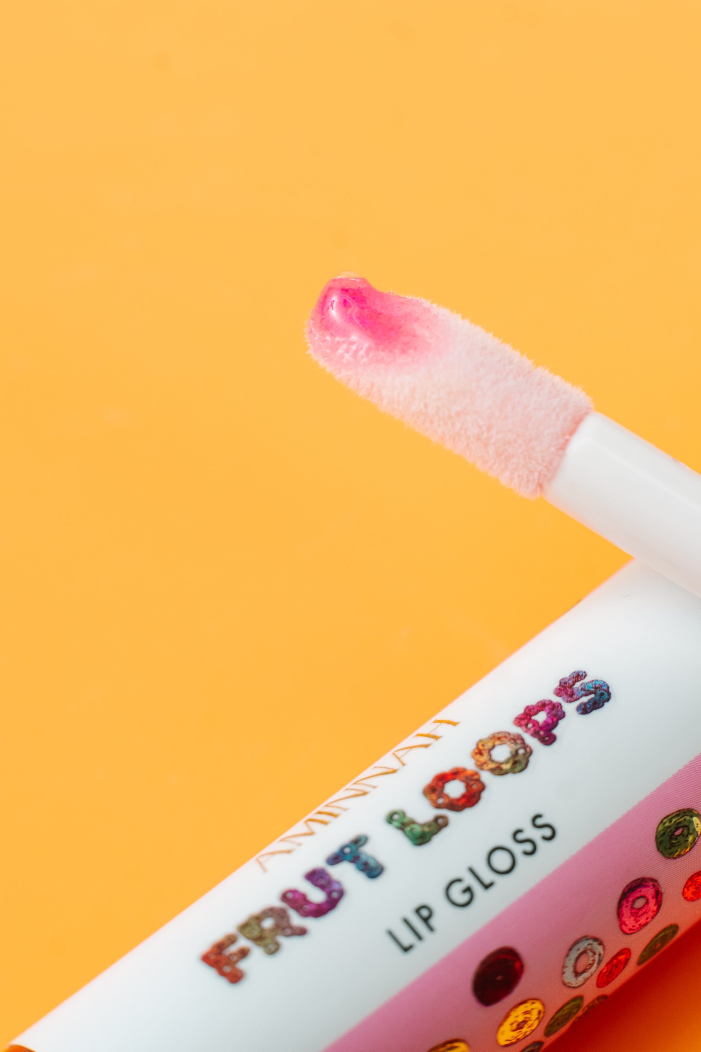 Close-up of the AMINNAH Frut Loops Lip Gloss doe-foot applicator with glossy pink lip gloss on a warm peach background. Moisturizing vegan lip gloss made with coconut oil and naturally derived ingredients for soft, smooth, high-shine lips. Clean beauty, cruelty-free, female-owned brand, made in the USA, available at Tree of Life Grocers.