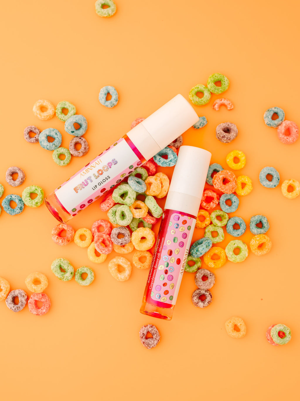 Two tubes of AMINNAH Frut Loops Lip Gloss surrounded by colorful fruity cereal pieces on a peach background. Moisturizing high-shine vegan lip gloss made with coconut oil and naturally derived ingredients for soft, hydrated lips. Clean beauty, cruelty-free, female-owned brand, made in the USA, available at Tree of Life Grocers.