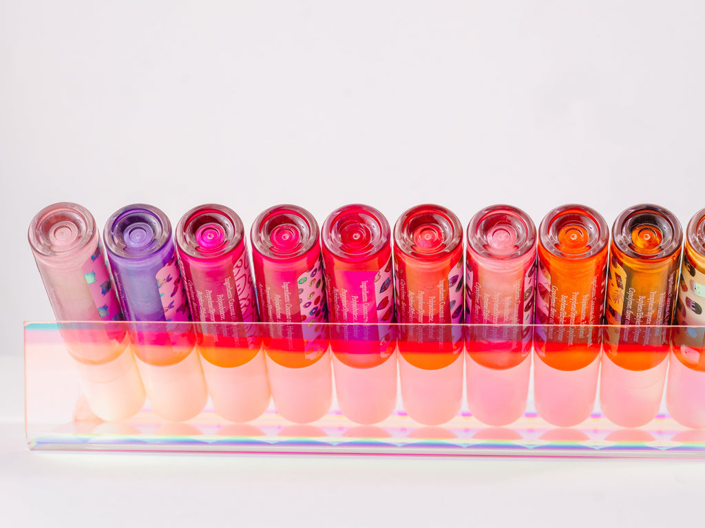 Collection of AMINNAH Frut Loops Lip Gloss shades displayed in a clear acrylic holder, showcasing a variety of vibrant pink, coral, orange, nude, and berry glosses. High-shine moisturizing vegan lip gloss made with coconut oil and naturally derived ingredients. Clean beauty, cruelty-free, female-owned brand, made in the USA, available at Tree of Life Grocers.