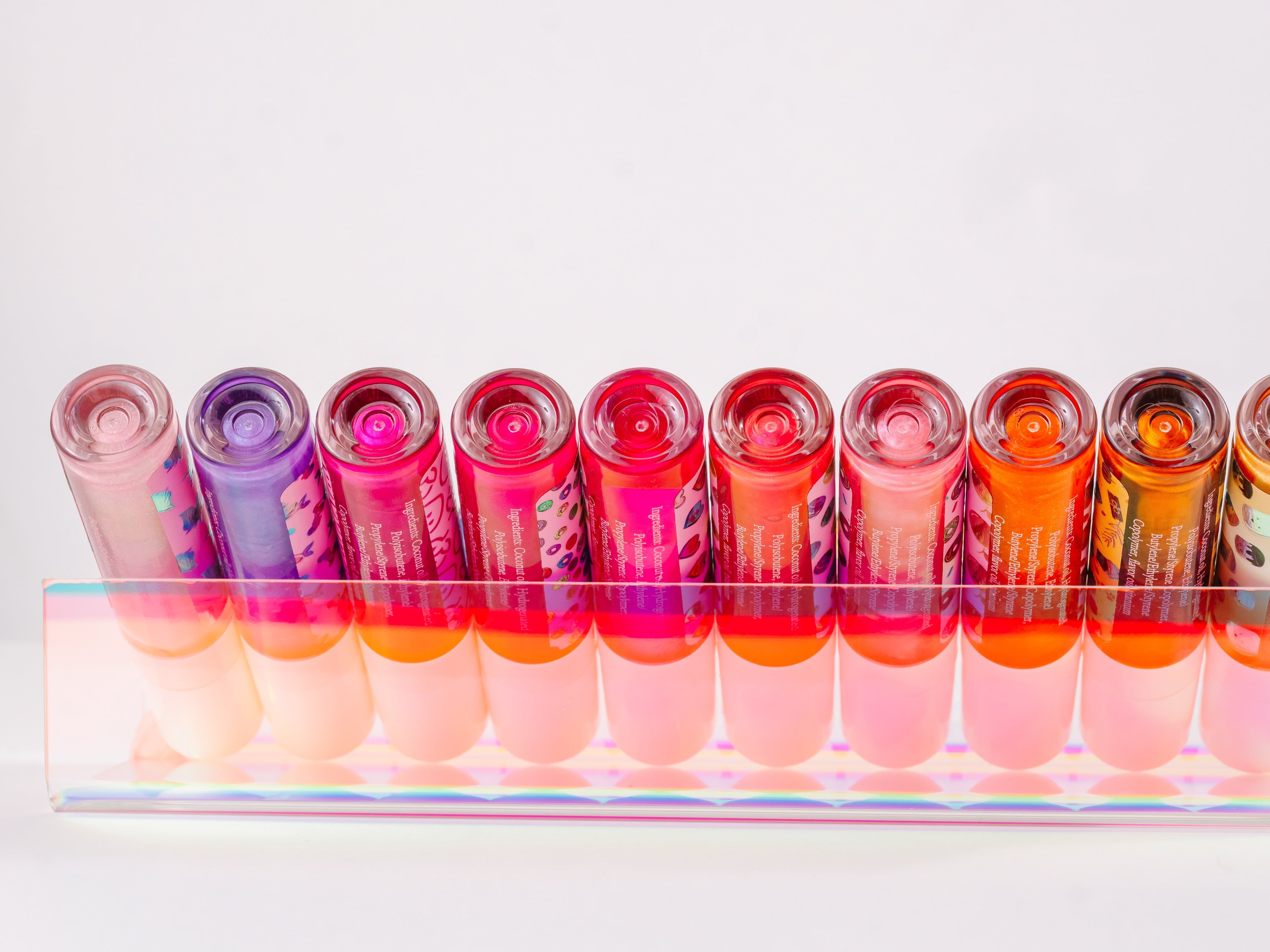 Collection of AMINNAH Frut Loops Lip Gloss shades displayed in a clear acrylic holder, showcasing a variety of vibrant pink, coral, orange, nude, and berry glosses. High-shine moisturizing vegan lip gloss made with coconut oil and naturally derived ingredients. Clean beauty, cruelty-free, female-owned brand, made in the USA, available at Tree of Life Grocers.