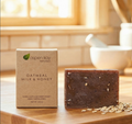 A bar of oatmeal milk and honey exfoliating soap stands next to its kraft paper packaging box on a wooden surface, with scattered oats and a softly blurred kitchen background.