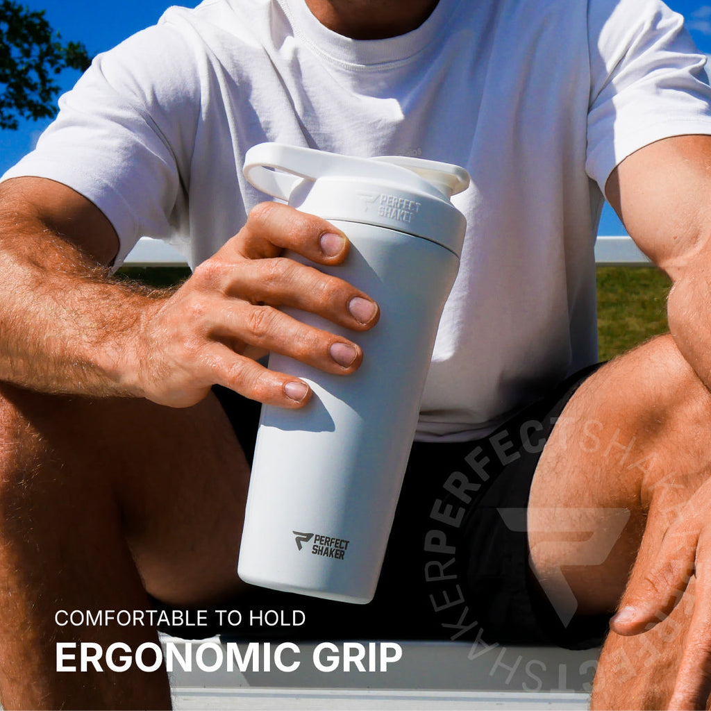 A person outdoors holding a white Perfect Shaker shaker bottle with an ergonomic grip. The person is wearing a white t-shirt and black shorts, with blue sky and grass in the background.