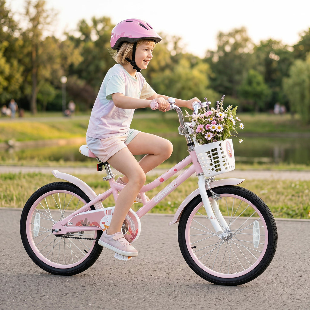 A young child wearing a pink helmet rides a pink children's bicycle with a white front basket filled with flowers on a paved path in a park at sunset.