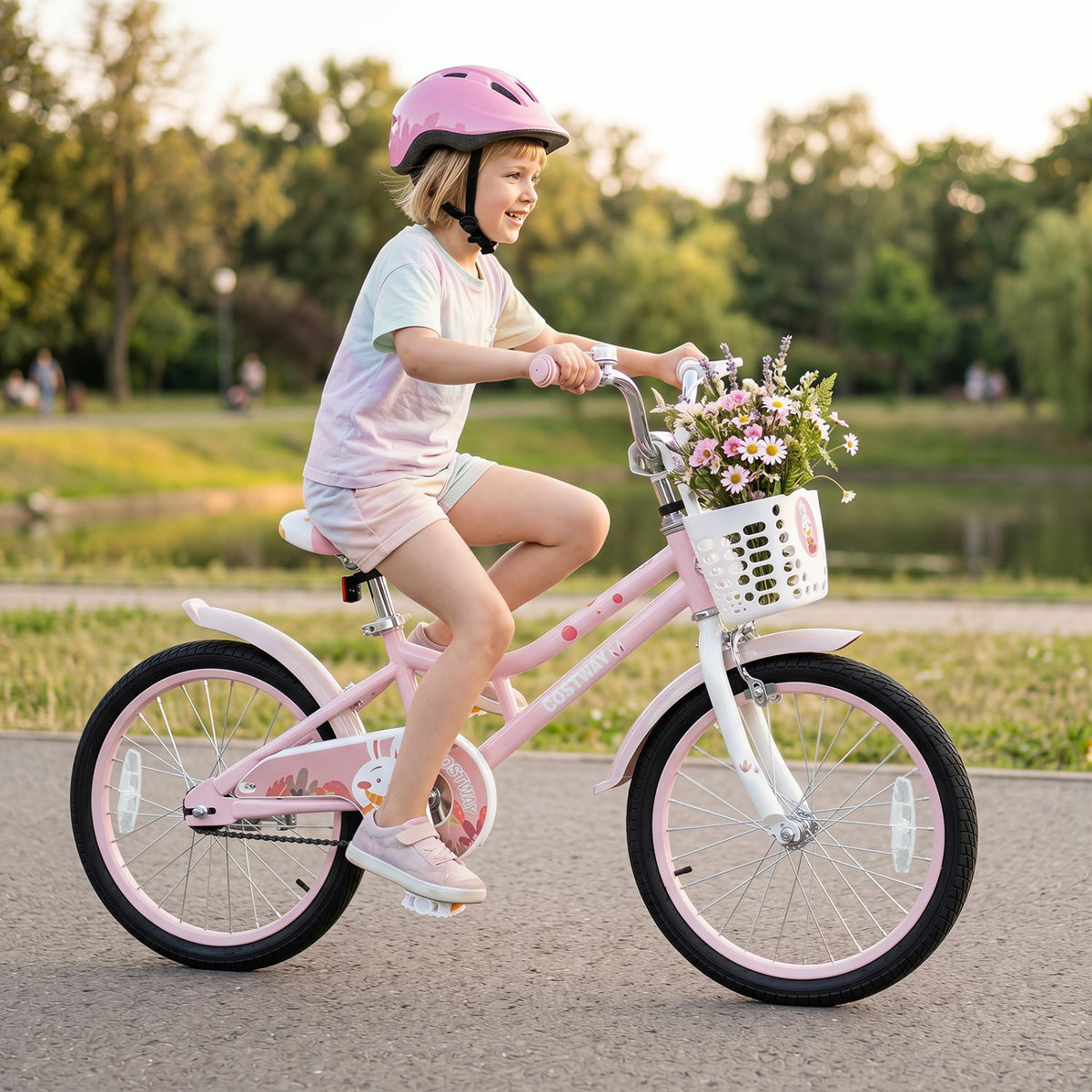 A young child wearing a pink helmet rides a pink children's bicycle with a white front basket filled with flowers on a paved path in a park at sunset.