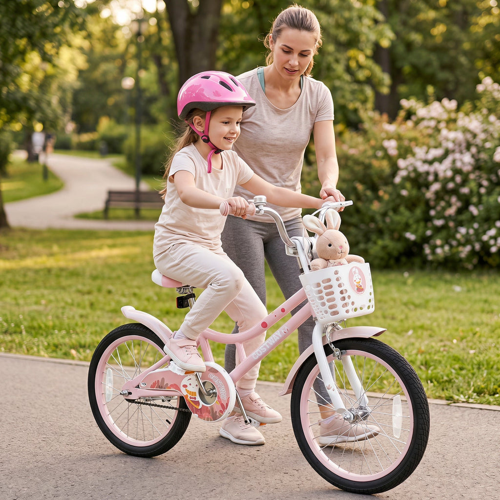 A young girl wearing a pink helmet rides a pink children's bicycle while her mother pushes her along a paved path in a park. The bicycle has a white basket with a stuffed bunny and a small cartoon character attached.