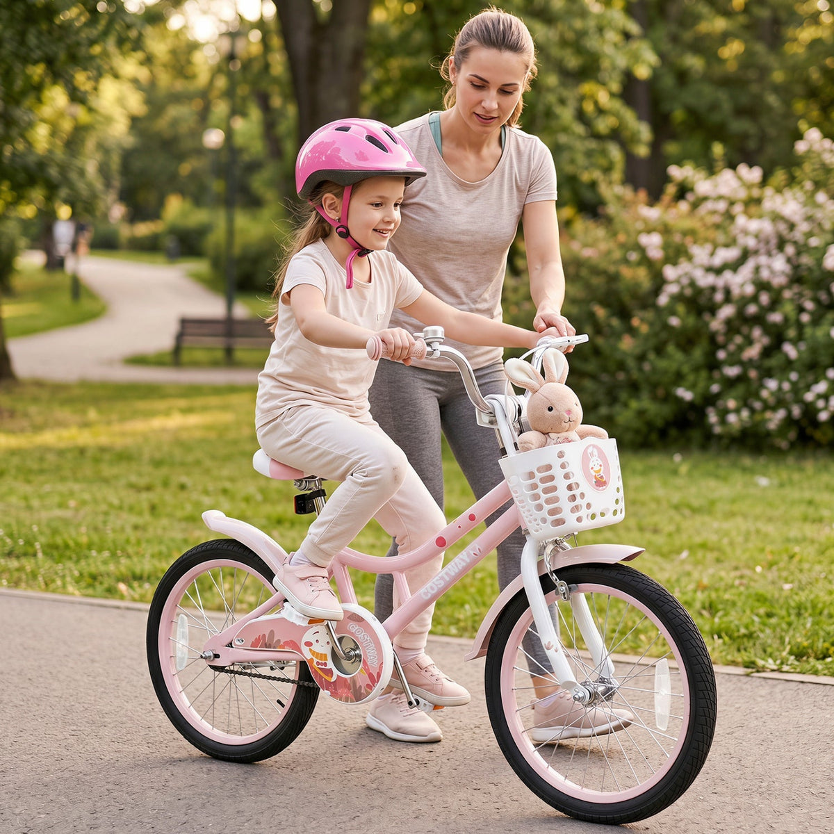 A young girl wearing a pink helmet rides a pink children's bicycle while her mother pushes her along a paved path in a park. The bicycle has a white basket with a stuffed bunny and a small cartoon character attached.