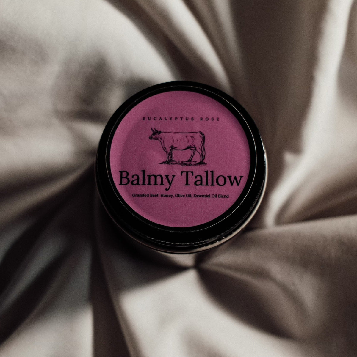 A round black jar lid with a pink label reading 'Eucalyptus Rose' and 'Balmy Tallow' with a cow illustration, placed on draped satin fabric. Eucalyptus Rose Tallow Balm 4 oz. | Nourishing Floral Skincare | Balmy Tallow Grassfed beef, honey, olive oil, essential oil blend.