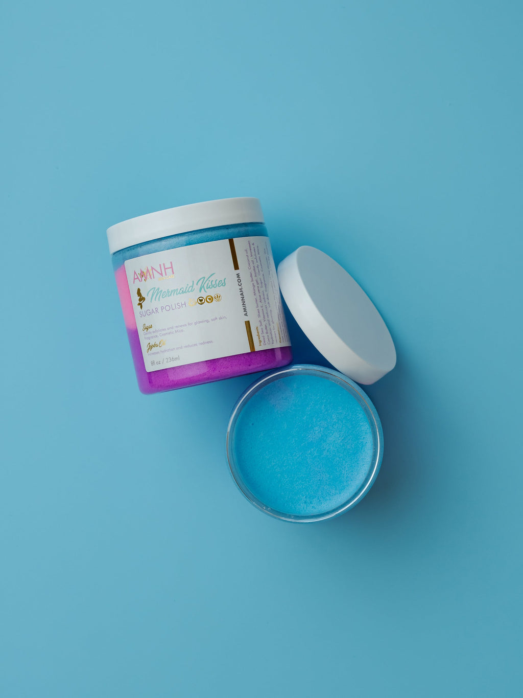 Open jar of blue powder with a purple label on a blue background. Mermaid Kisses Sugar Scrub with shimmering blue natural sugar scrub in an open jar and labeled container on a soft blue background. Clean beauty, cruelty-free, vegan body exfoliator made with natural ingredients by a female-owned brand for smoother, softer, glowing skin.