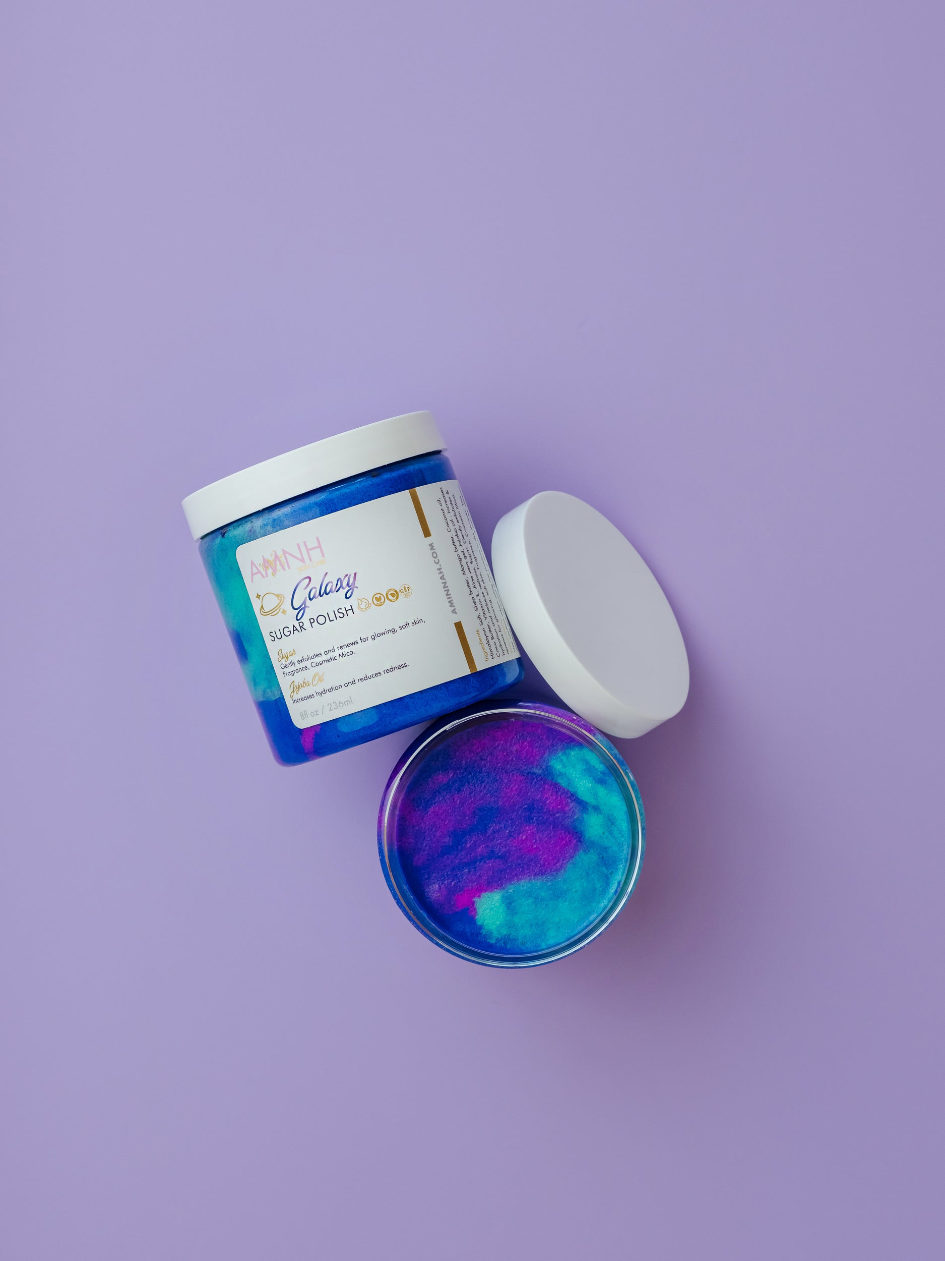 AMINNAH Galaxy Sugar Scrub in a jar with vibrant blue and purple sugar crystals on a lavender background. Clean beauty, cruelty-free, vegan body scrub made with natural ingredients to gently exfoliate, hydrate, smooth, and reveal glowing skin. Female-owned brand available at Tree of Life Grocers.