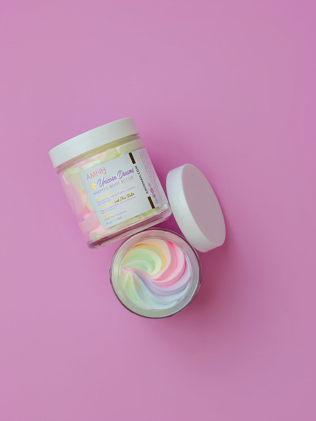 Unicorn Dreams Whipped Body Butter in an open glass jar with pastel rainbow whipped body butter beside its matching container on a pink background. Made with natural ingredients including shea butter, cocoa butter, mango butter, coconut oil, jojoba oil, and vitamin E. Clean beauty, cruelty-free, vegan, female-owned brand that deeply moisturizes, softens, and nourishes dry skin with a sweet cotton candy fragrance.