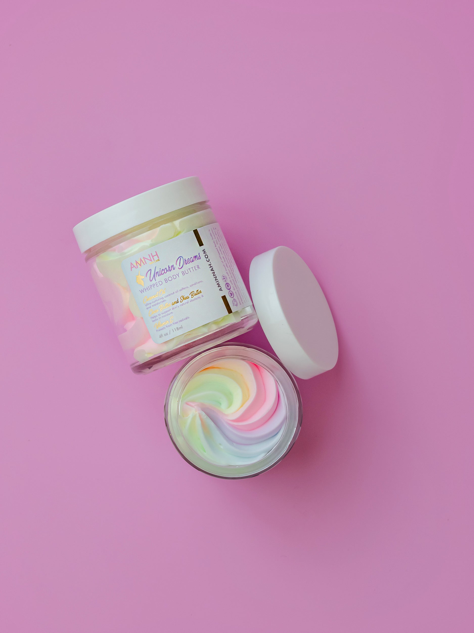 Unicorn Dreams Whipped Body Butter in an open glass jar with pastel rainbow whipped body butter beside its matching container on a pink background. Made with natural ingredients including shea butter, cocoa butter, mango butter, coconut oil, jojoba oil, and vitamin E. Clean beauty, cruelty-free, vegan, female-owned brand that deeply moisturizes, softens, and nourishes dry skin with a sweet cotton candy fragrance.