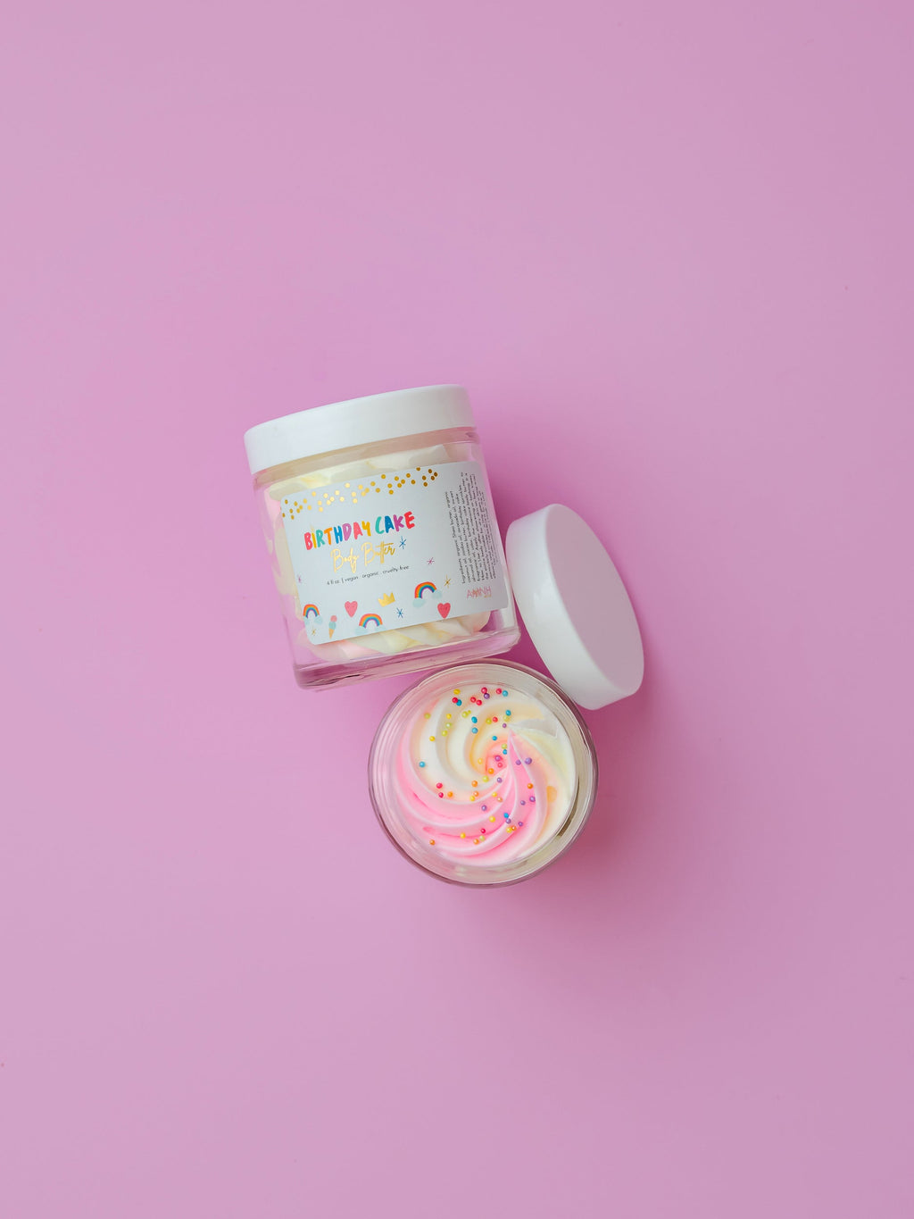 AMINNAH Birthday Cake Whipped Body Butter closed and open jars displayed on a pink background, showcasing the creamy whipped texture with colorful sprinkles. Moisturizing body butter made with natural ingredients including shea butter, cocoa butter, mango butter, coconut oil, jojoba oil, and vitamin E. Clean Beauty, cruelty-free, vegan, female-owned brand skincare proudly made in the USA, available at Tree of Life Grocers.