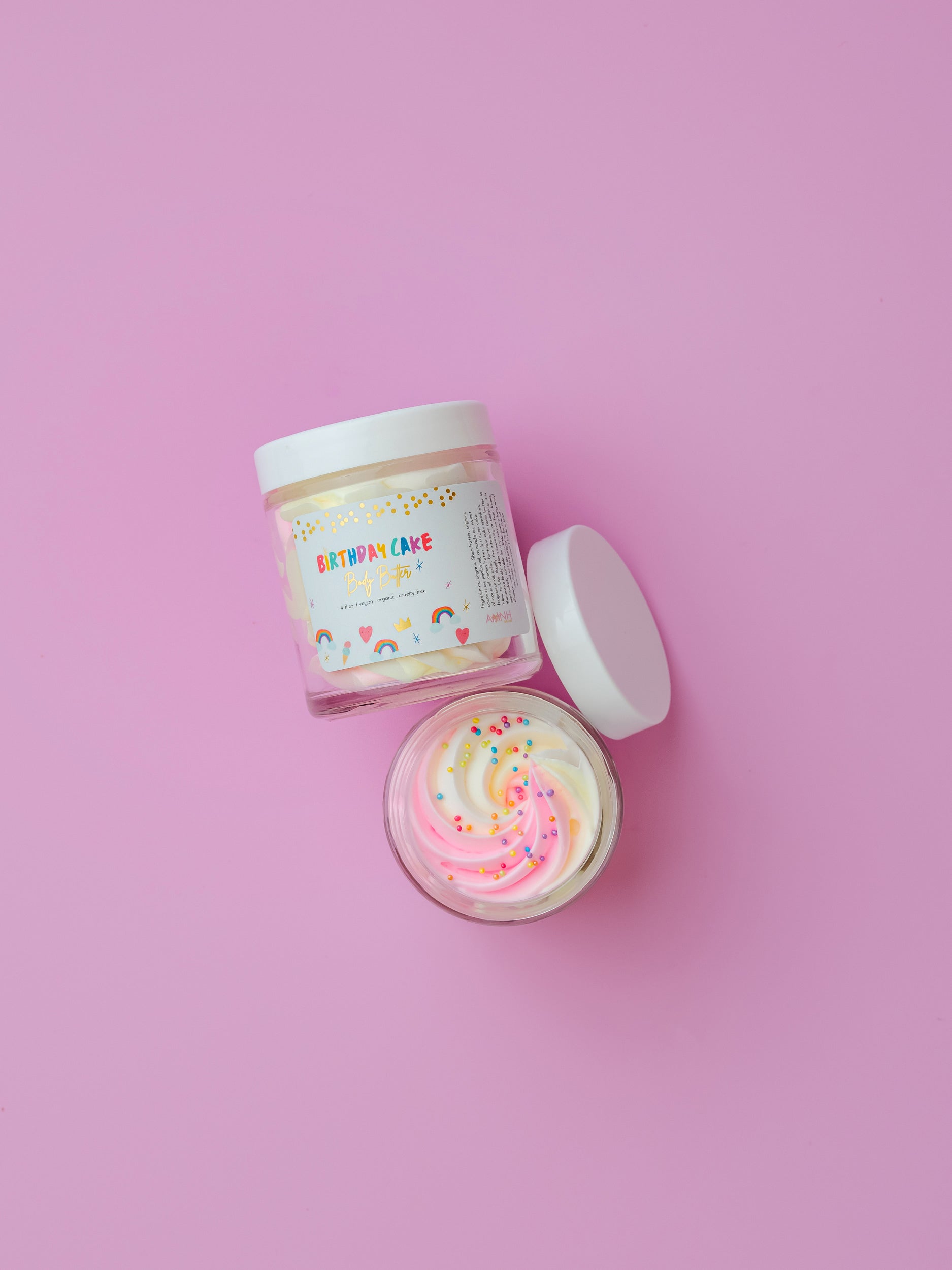AMINNAH Birthday Cake Whipped Body Butter closed and open jars displayed on a pink background, showcasing the creamy whipped texture with colorful sprinkles. Moisturizing body butter made with natural ingredients including shea butter, cocoa butter, mango butter, coconut oil, jojoba oil, and vitamin E. Clean Beauty, cruelty-free, vegan, female-owned brand skincare proudly made in the USA, available at Tree of Life Grocers.