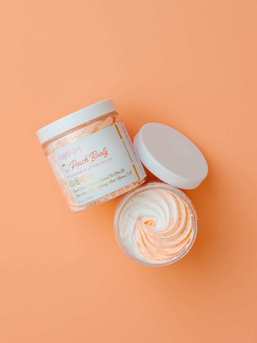 Open jar of body scrub with a decorative label on an orange background. Open jar of AMINNAH Peach Booty Tightening & Lifting Scrub with its creamy whipped exfoliating texture beside the product jar on a peach background. Clean beauty, cruelty-free, vegan body scrub made with natural ingredients including Peach Extract, Aloe Vera, Retinol, and Apricot Seed Powder from a female-owned brand.