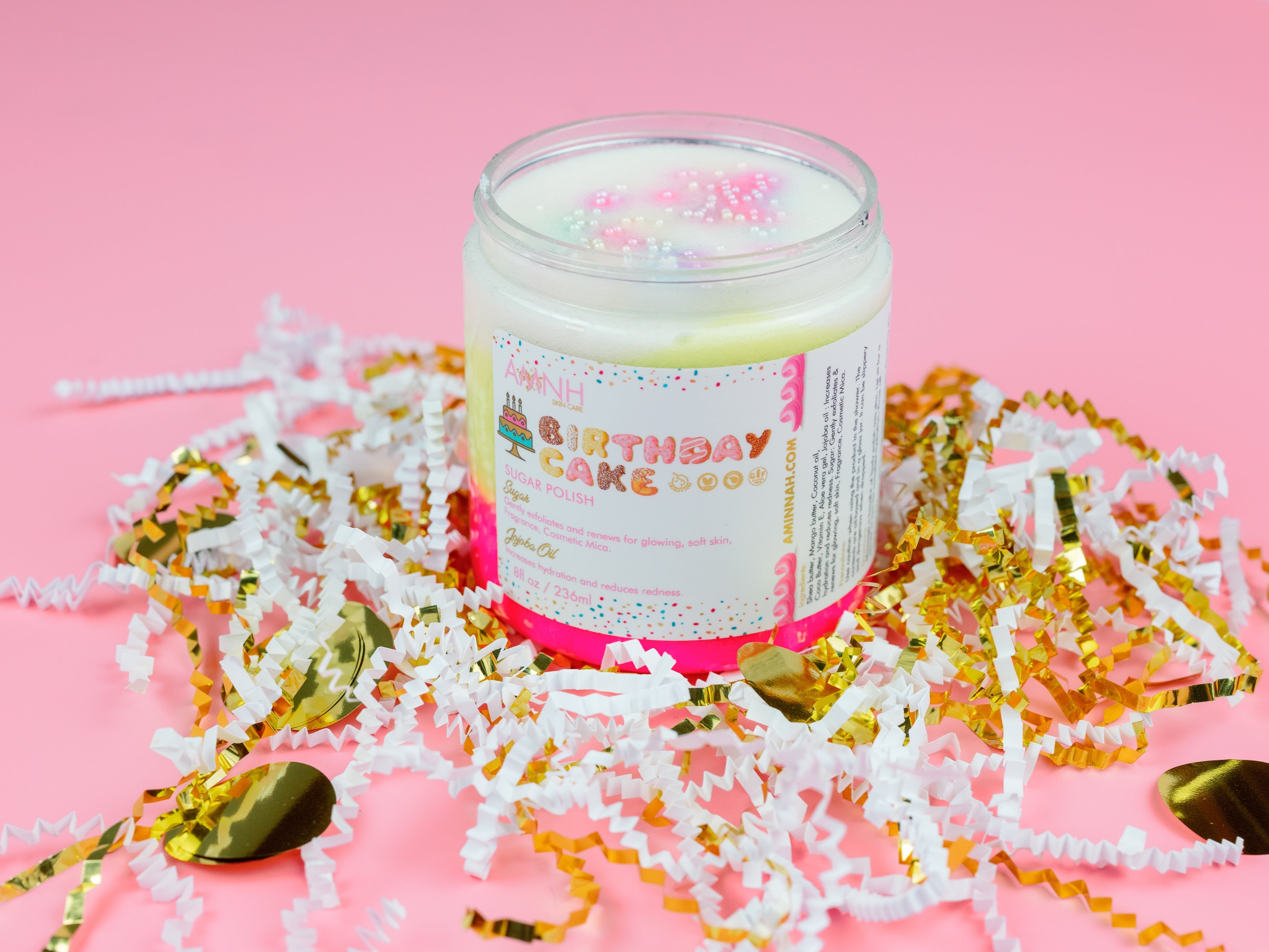 Birthday Cake Sugar Scrub in a reusable jar surrounded by festive gold and white confetti on a soft pink background. This clean beauty, cruelty-free, vegan sugar scrub is handcrafted with natural ingredients by a female-owned brand to gently exfoliate, deeply hydrate, and leave skin soft, smooth, and glowing with a delicious birthday cake-inspired scent.