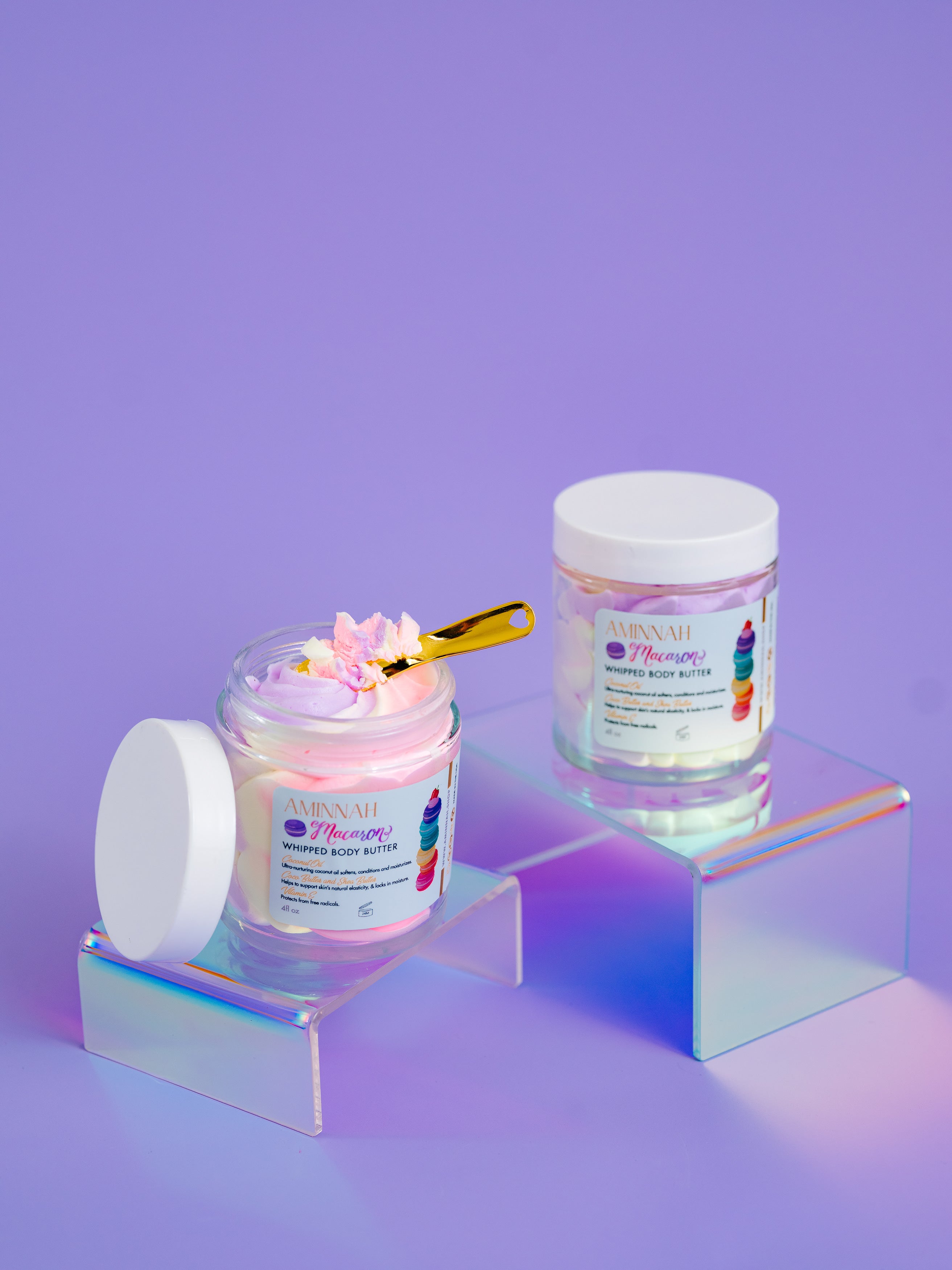 Two jars of AMINNAH Macaron Whipped Body Butter displayed on iridescent acrylic risers against a soft lavender background, with one open jar revealing a fluffy pink whipped texture and a gold spatula. Inspired by the sweet scent of French macarons, this luxurious body butter is crafted with nourishing botanical butters and plant oils to deeply hydrate, soften, and smooth dry skin. Clean beauty, cruelty-free, vegan, and made with naturally inspired ingredients by a female-owned brand. At Tree of Life Grocers