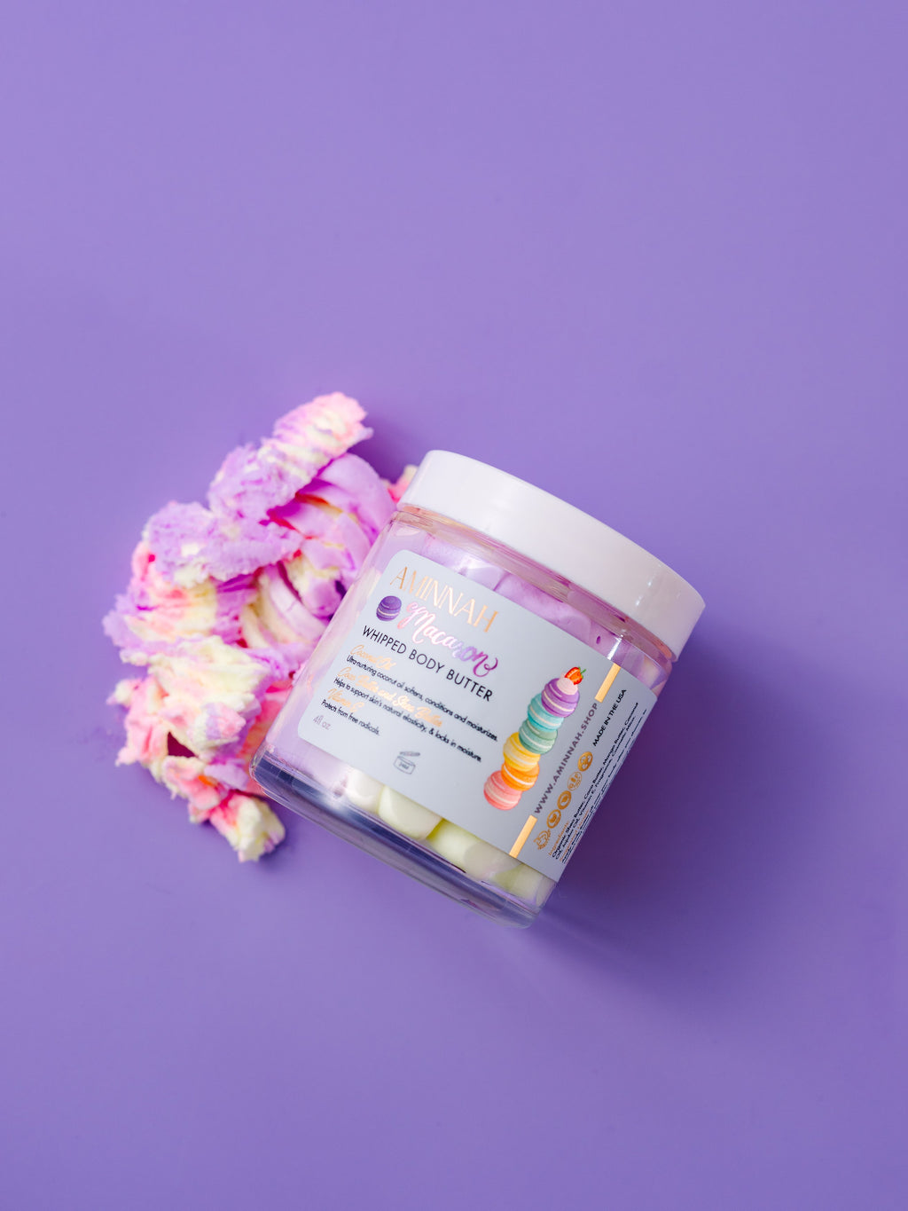 AMINNAH Macaron Whipped Body Butter jar displayed on a soft lavender background beside pastel macaron-inspired decorative accents. This luxurious whipped body butter is inspired by the sweet scent of French macarons and is crafted with nourishing botanical butters and plant oils to deeply hydrate, soften, and smooth dry skin. Clean beauty, cruelty-free, vegan, and made with naturally inspired ingredients by a female-owned brand. Available at Tree of Life Grocers.