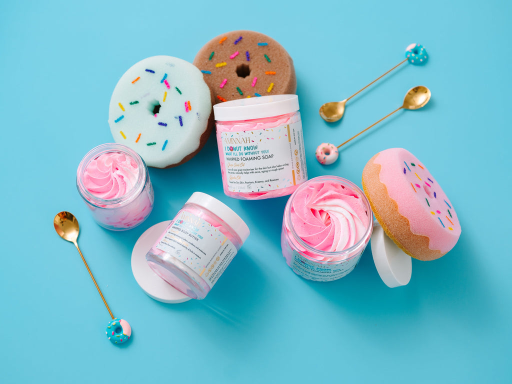 I Donut Know What I'll Do Without You! Whipped Body Butter in a playful donut-inspired display featuring fluffy pink whipped body butter in open jars, decorative sprinkle donuts, and gold spoons. Richly formulated with shea butter, cocoa butter, mango butter, coconut oil, jojoba oil, vitamin E, and fruit extracts to deeply moisturize, soften, and nourish dry skin. Cruelty-free, made with clean ingredients, and perfect for daily hydration.