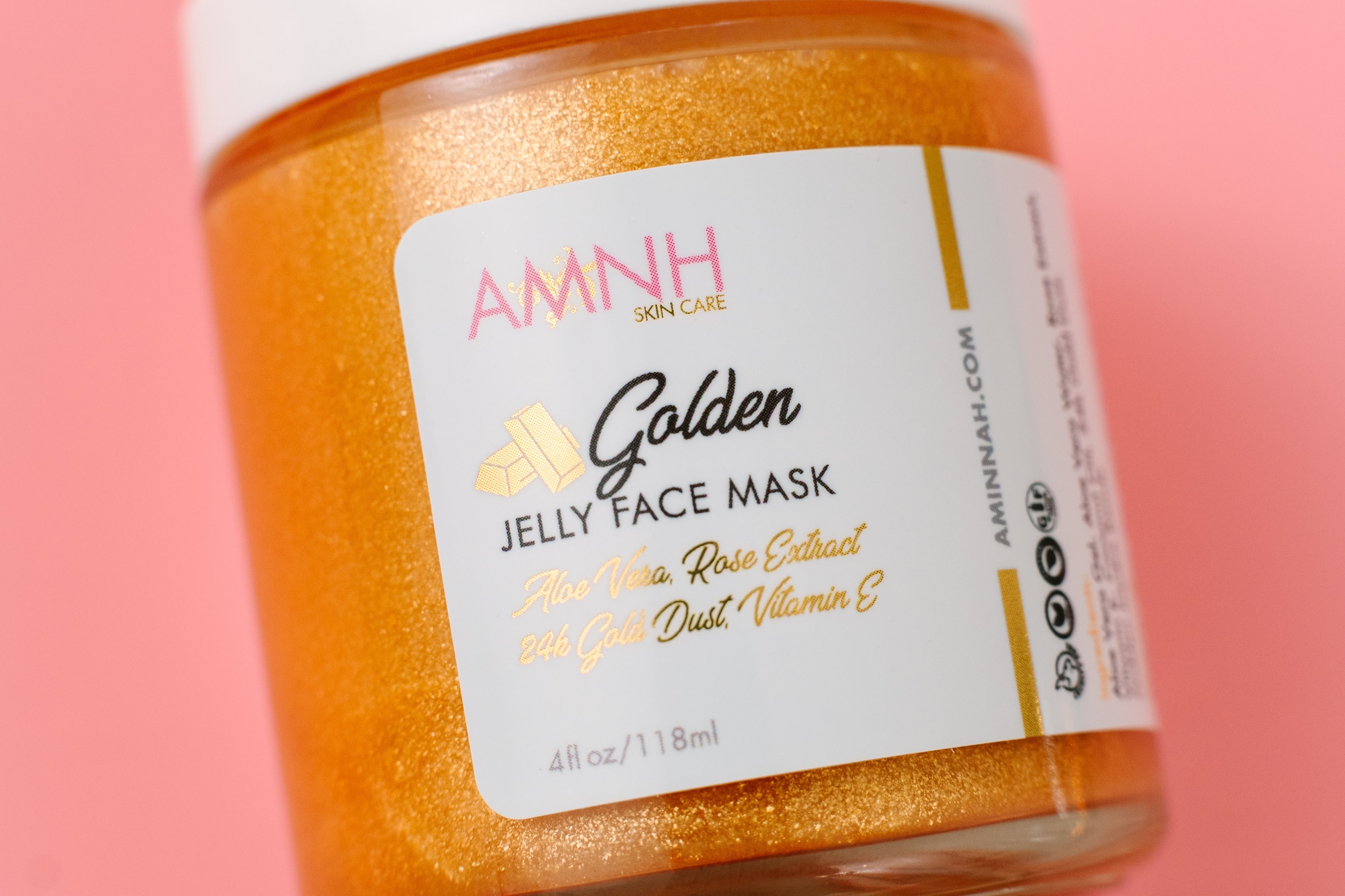 24K Golden Jelly Face Mask in a glass jar with shimmering gold formula on a pink background. Clean beauty, cruelty-free, vegan skincare made with natural ingredients including aloe vera, rose extract, vitamin E, coconut oil, 24K gold flakes, and jojoba beads. Female-owned brand designed to hydrate, soothe, firm, brighten, reduce the appearance of fine lines, and support anti-aging for healthy, radiant, glowing skin.