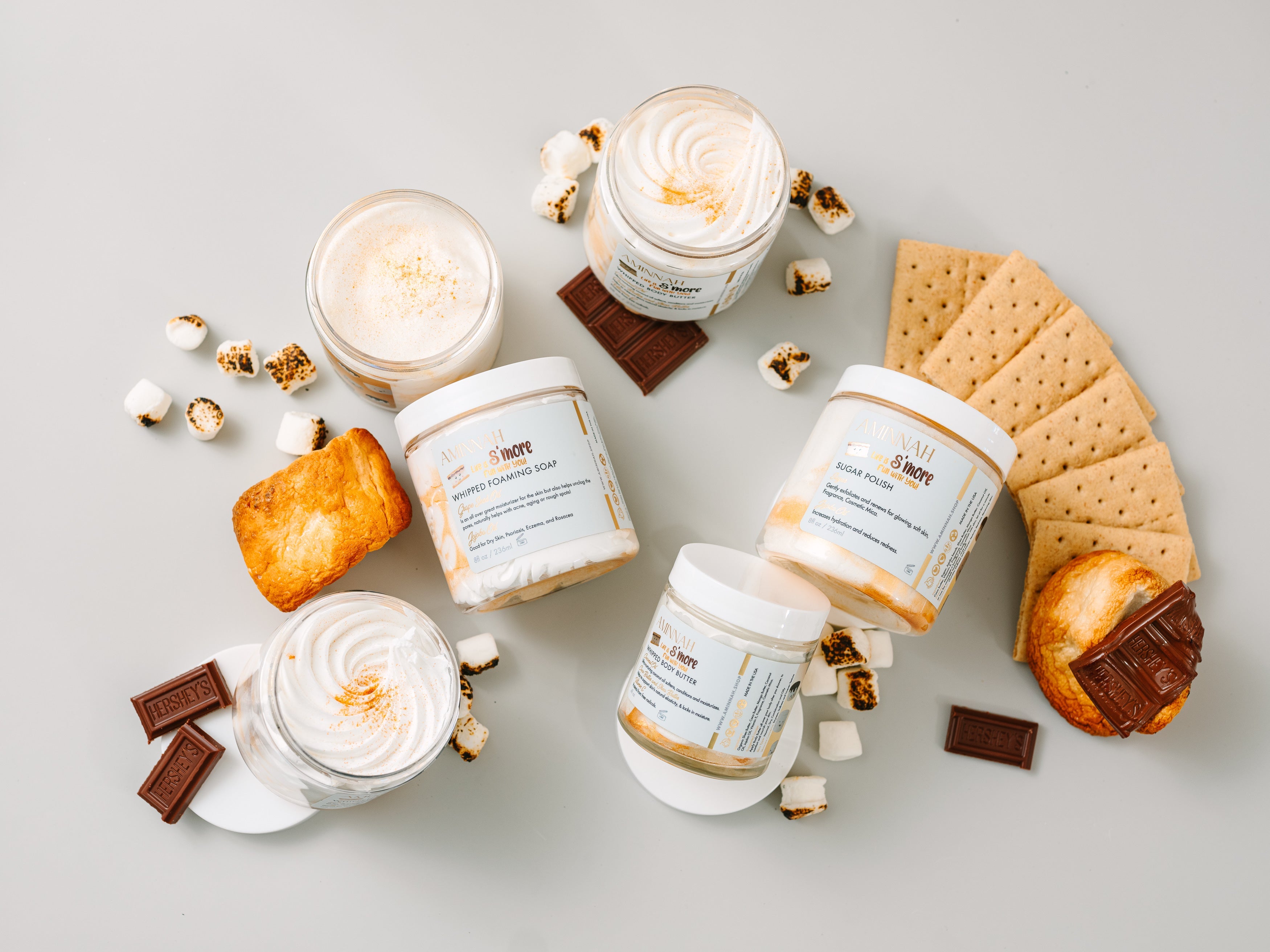 AMINNAH® Life is S’more Fun With You Sugar Scrub flat lay with toasted marshmallow, chocolate, graham crackers, and whipped sugar scrub. Clean beauty, cruelty-free, vegan, paraben-free, gluten-free skincare made with natural ingredients in recyclable packaging. Female-owned brand body exfoliator with shea butter, cocoa butter, coconut oil, jojoba oil, aloe vera, vitamin E, and 24K gold for soft, smooth, hydrated skin.