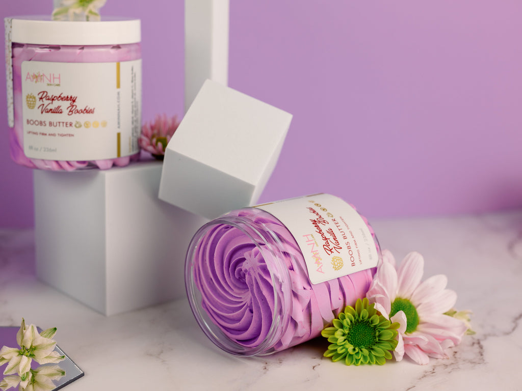 AMINNAH "My Iced Gems" Boob Butter in Raspberry Vanilla displayed in decorative jars with whipped lavender body butter, white display cubes, and fresh flowers on a soft purple background. Vegan, cruelty-free body butter for nourishing and moisturizing skin. Clean beauty, cruelty-free, vegan, made with naturally derived ingredients, and crafted by a female-owned brand. Available at Tree of Life Grocers.