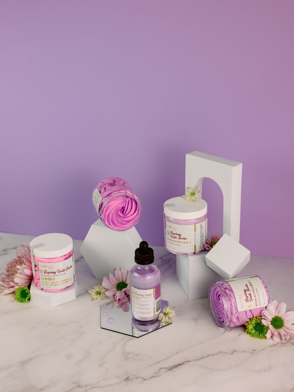 AMINNAH "My Iced Gems" Boob Butter skincare collection featuring whipped body butter jars, body oil, and pastel purple spa products arranged with flowers on a marble surface. Clean beauty, cruelty-free, vegan, made with naturally derived ingredients, and crafted by a female-owned brand. Available at Tree of Life Grocers.