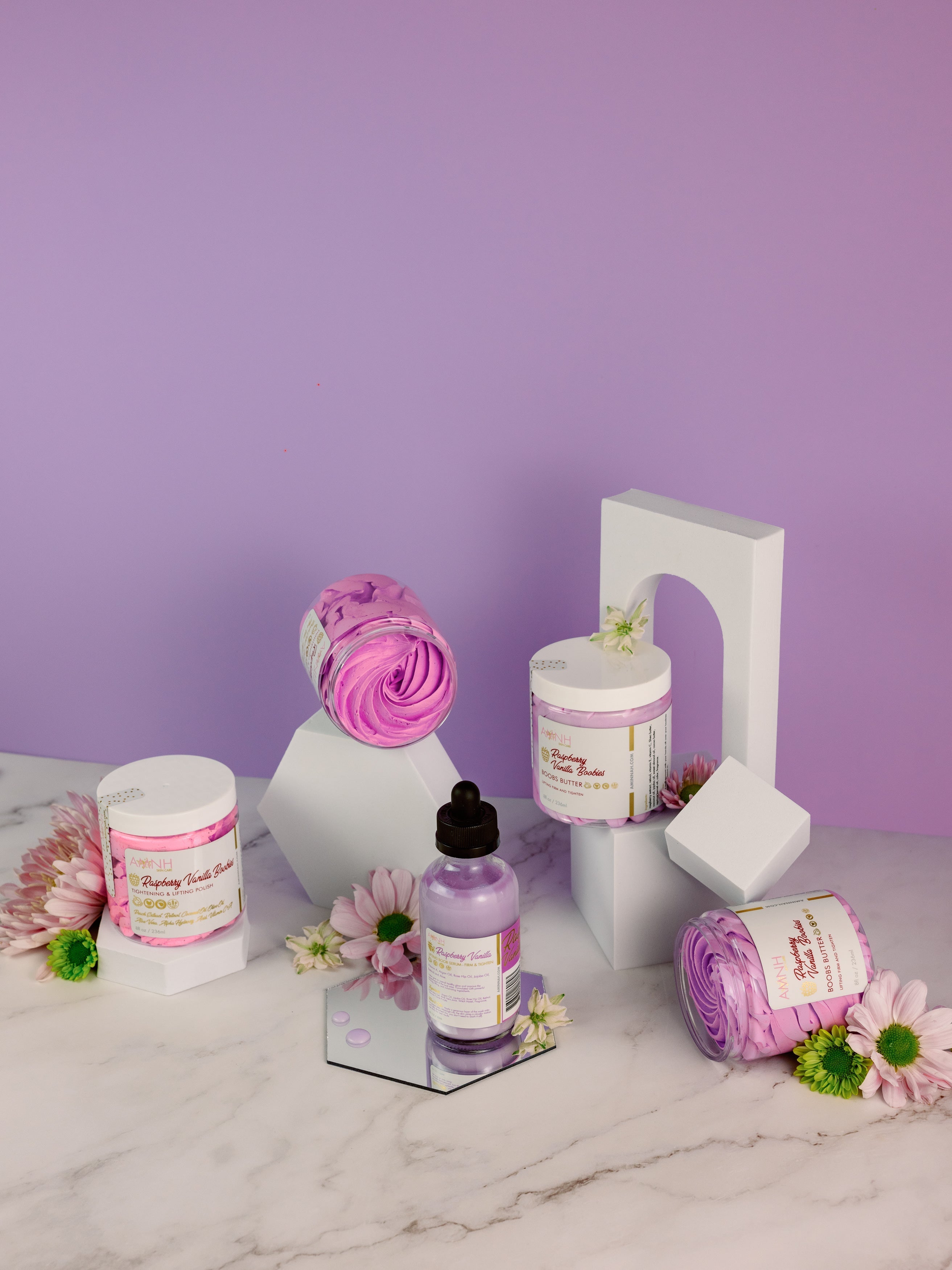 AMINNAH "My Iced Gems" Boob Butter skincare collection featuring whipped body butter jars, body oil, and pastel purple spa products arranged with flowers on a marble surface. Clean beauty, cruelty-free, vegan, made with naturally derived ingredients, and crafted by a female-owned brand. Available at Tree of Life Grocers.