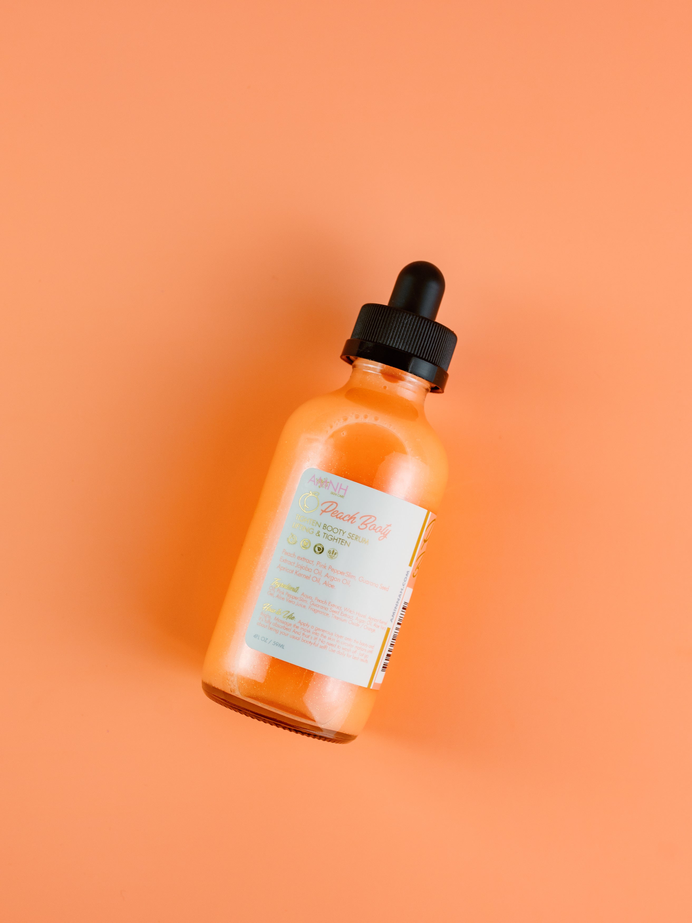 AMINNAH Peach Booty Butt Serum in amber glass dropper bottle on a soft peach background. Clean beauty body serum made with natural ingredients to help hydrate, nourish, smooth, firm, and improve the appearance of skin. Cruelty-free, vegan skincare from a female-owned brand, perfect for luxurious booty care and daily self-care routines.