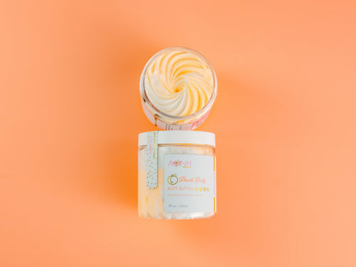 Open and closed jars of AMINNAH "Peach Booty" Tightening & Lifting Butt Butter displayed against a soft peach background, showcasing the rich whipped texture and included applicator spatula. This luxurious body butter is formulated with peach extract, retinol, vitamin C, vitamin E, shea butter, cocoa butter, coconut oil, jojoba oil, sweet almond oil, and apricot kernel oil to deeply hydrate, nourish, smooth, and improve the appearance of firmer-looking skin. Clean beauty, cruelty-free, vegan