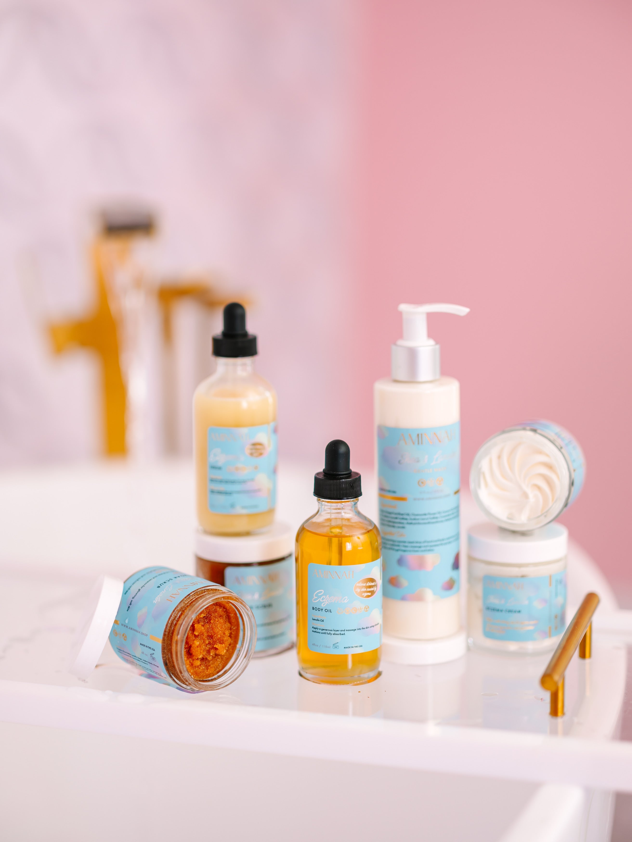 Natural skincare collection displayed on a bathroom sink featuring eczema cream, serum, sugar scrub, body oil, cleanser, and lotion in clean beauty packaging from a cruelty-free, vegan, female-owned brand.