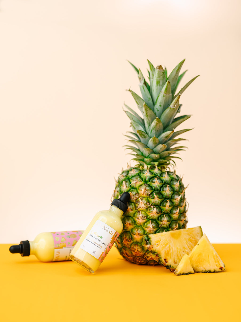 AMINNAH Fineapples Boob Serum bottles displayed beside a fresh pineapple and pineapple slices on a bright yellow background. Hydrating body and boob serum made with pineapple fruit extract, retinol, vitamin E, argan oil, jojoba oil, rosehip oil, aloe vera, and botanical oils to nourish, smooth, and soften skin. Clean beauty, cruelty-free, vegan, formulated with natural ingredients, and crafted by a female-owned brand for radiant, healthy-looking skin.