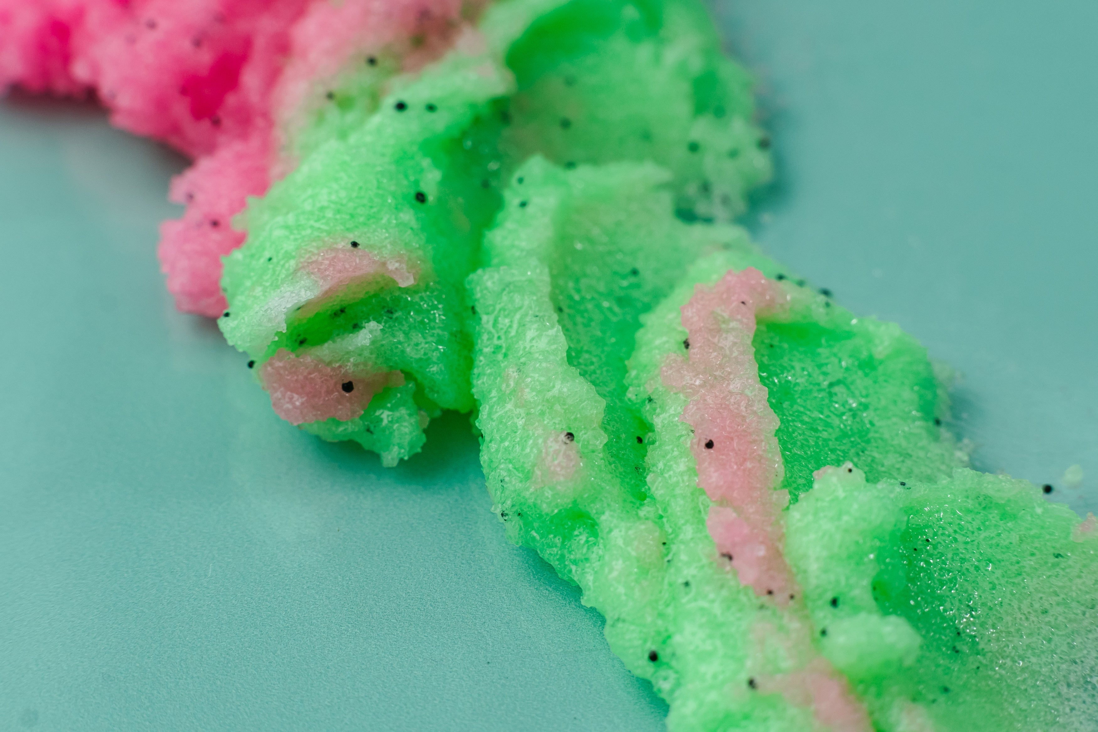 Close-up of green and pink textured material on a light blue background. Water My Melons Boob Sugar Scrub with exfoliating pink sugar scrub and branded container on a teal background. Clean beauty skincare made with natural ingredients including watermelon fruit extract, shea butter, cocoa butter, mango butter, aloe vera, vitamin E, retinol, and botanical oils. Cruelty-free, vegan, female-owned beauty brand.