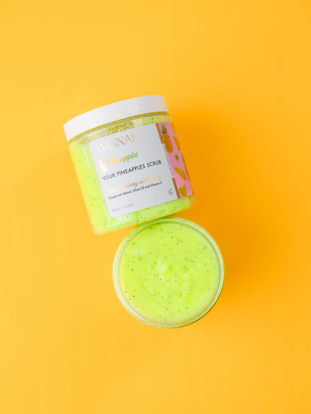 AMINNAH Fineapples Boob Sugar Scrub with pineapple, retinol, and vitamin E in an open jar on a yellow background. Luxury clean beauty body scrub made with natural ingredients to gently exfoliate and smooth skin. Vegan, cruelty-free, paraben-free, gluten-free formula from a female-owned brand with recyclable packaging. Anti-aging skincare for soft, radiant-looking skin.