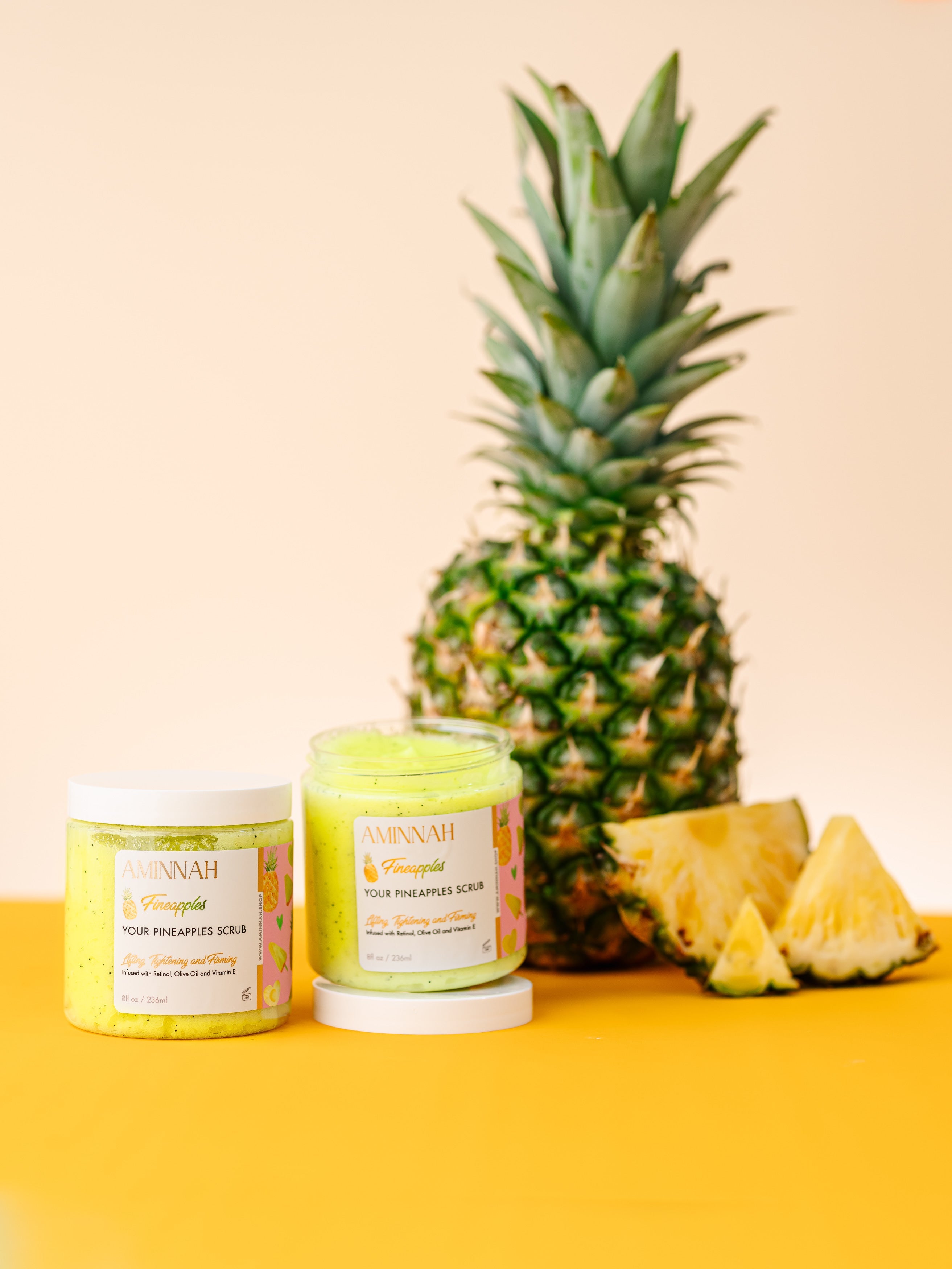 Fineapples Boob Sugar Scrub with pineapple, retinol and vitamin E in two jars beside a fresh pineapple on a yellow background. Anti-aging exfoliating body scrub with natural ingredients for smoother, radiant-looking skin. Clean beauty, cruelty-free, vegan, paraben-free, gluten-free, recyclable packaging, female-owned brand, luxurious body care.