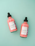 2 'Water My Melons' Boob Serum glass bottles on a green background. 'Water My Melons' Boob Serum by AMINNAH – hydrating breast serum with natural ingredients for soft, nourished skin. Clean beauty, cruelty-free skincare from a female-owned brand. Vegan formula made with skin-loving botanical ingredients for daily hydration and self-care.
