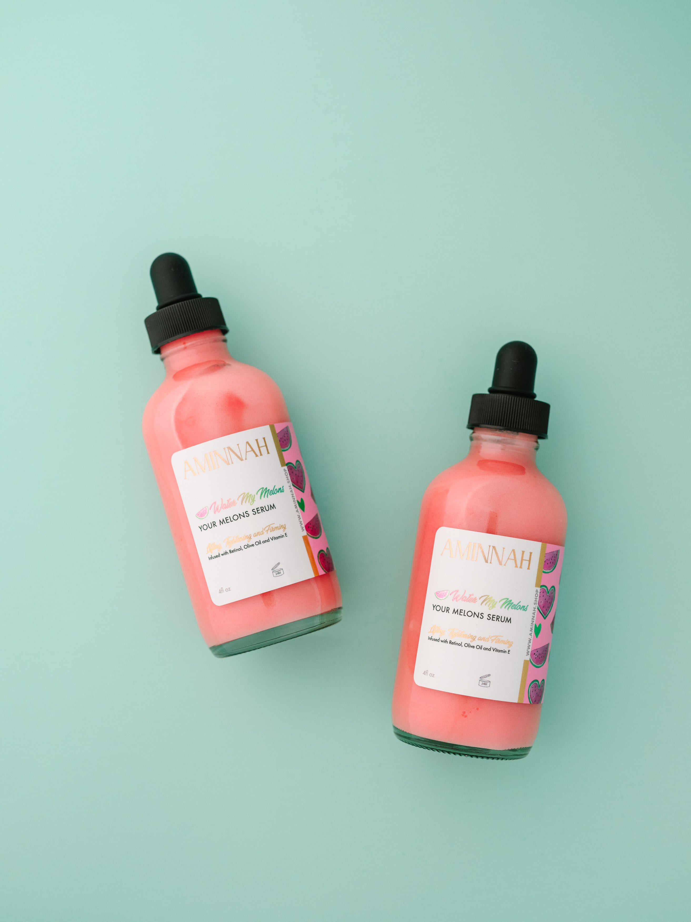 2 'Water My Melons' Boob Serum glass bottles on a green background. 'Water My Melons' Boob Serum by AMINNAH – hydrating breast serum with natural ingredients for soft, nourished skin. Clean beauty, cruelty-free skincare from a female-owned brand. Vegan formula made with skin-loving botanical ingredients for daily hydration and self-care.
