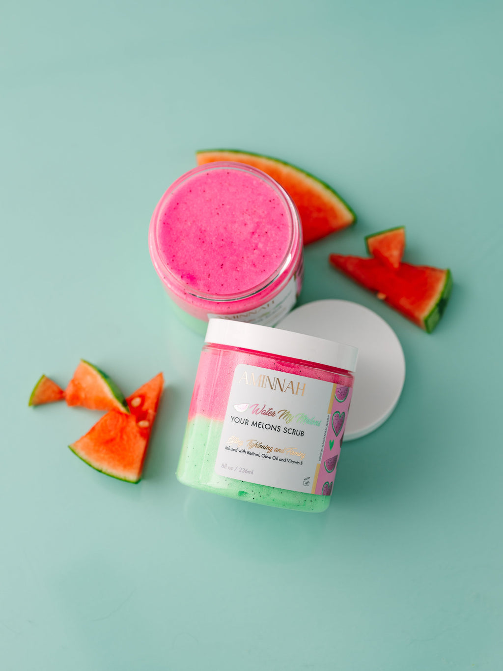 Pink and green jar of body scrub with watermelon slices on a light blue background. Water My Melons Boob Sugar Scrub in an open jar with exfoliating pink sugar scrub and branded container on a teal background. Clean beauty skincare made with natural ingredients including watermelon fruit extract, shea butter, cocoa butter, mango butter, aloe vera, vitamin E, retinol, and botanical oils. Cruelty-free, vegan, female-owned beauty brand.